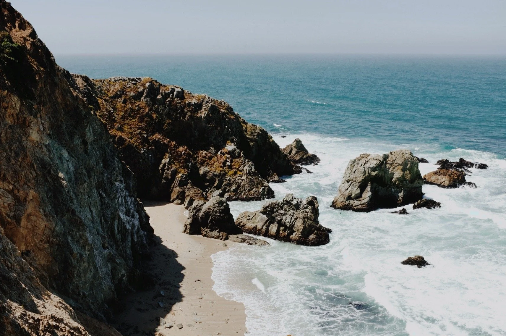 Rocky coastline with cliffs, large rocks in the ocean, and waves crashing on a sandy beach under a clear sky.