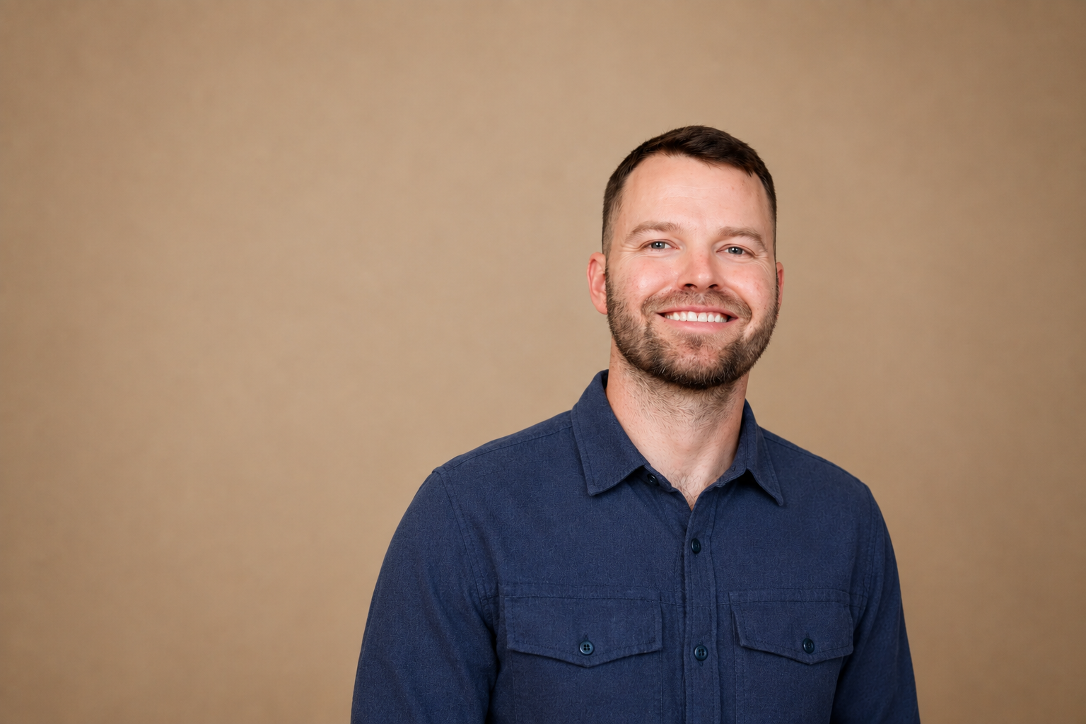 A smiling man with short brown hair, beard, and blue eyes, wearing a dark blue button-up shirt, standing against a plain beige background.