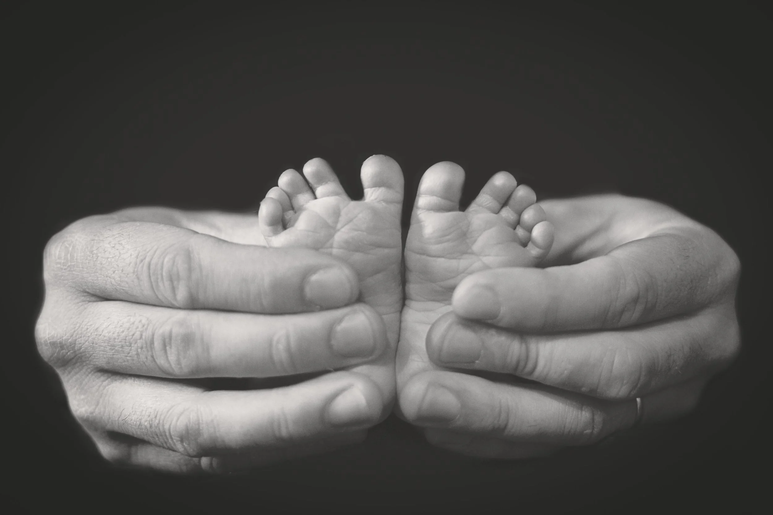 Black and white close-up of a newborn baby's feet held in a parent's hands — a tender detail captured at home in London by Kate See.