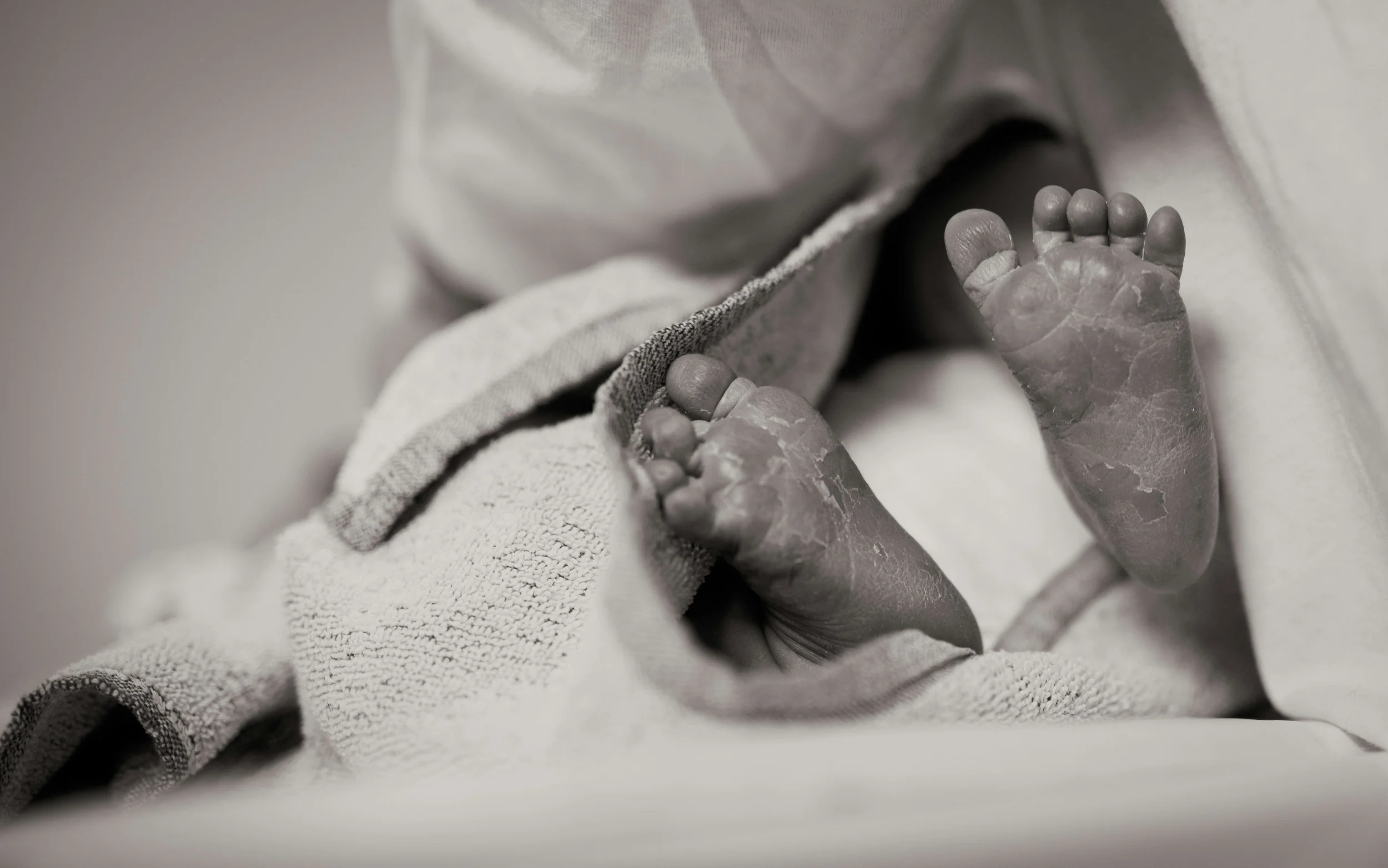 Close-up of a newborn baby's feet with tiny toes, wrapped in a soft cloth with a textured blanket.