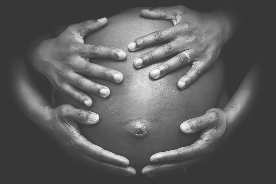 Black and white photograph of multiple hands forming a heart shape around a pregnant belly.