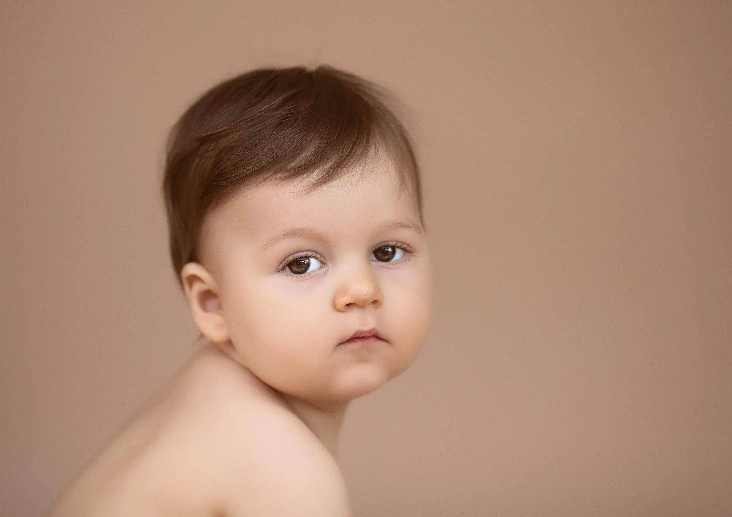 Close-up of a young child's face with short brown hair, brown eyes, and a neutral expression, set against a plain beige background.
