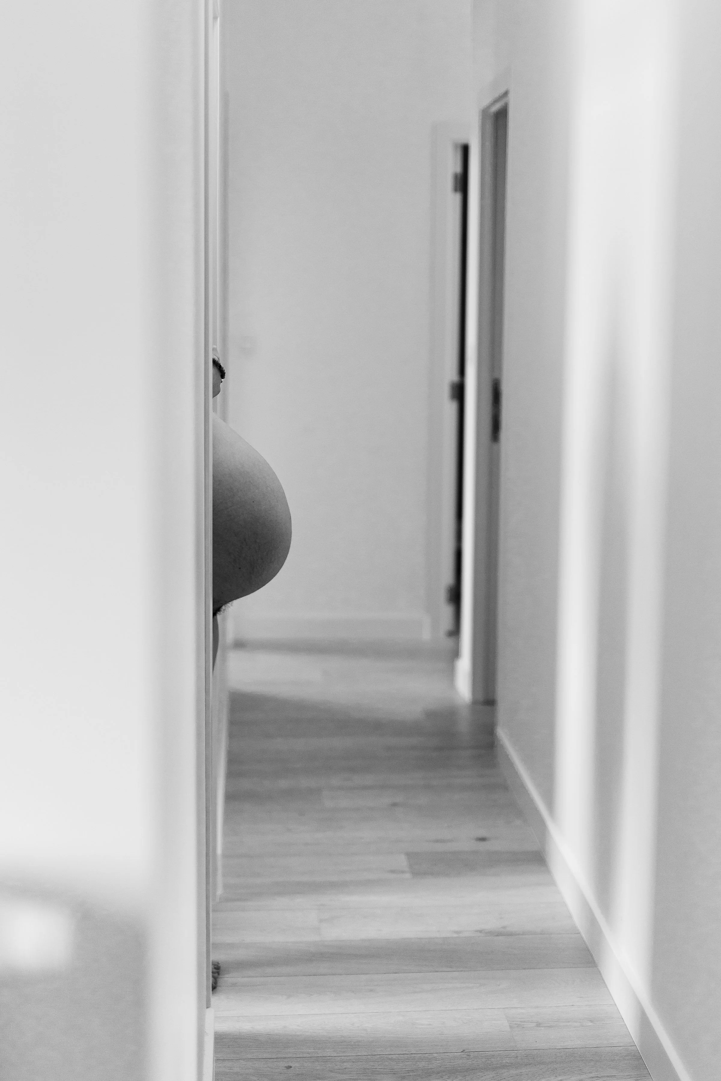 A black and white photo showing a hallway with a person partially visible, only their lower back and a part of their side. The person is standing near a wall, with the hallway extending towards a door or opening in the background.