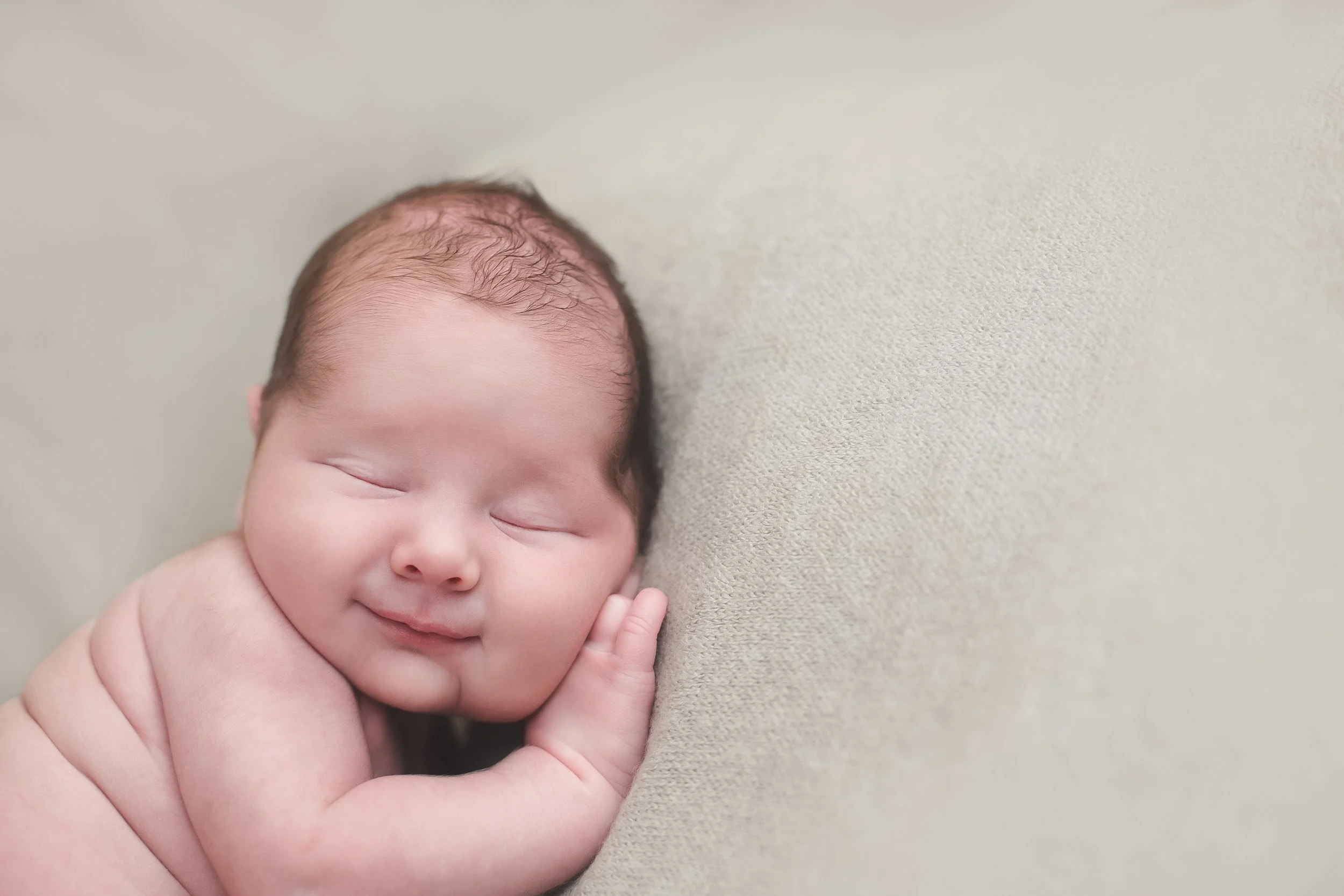 A peaceful sleeping newborn baby with closed eyes and a content expression, resting on a soft, neutral-colored surface.