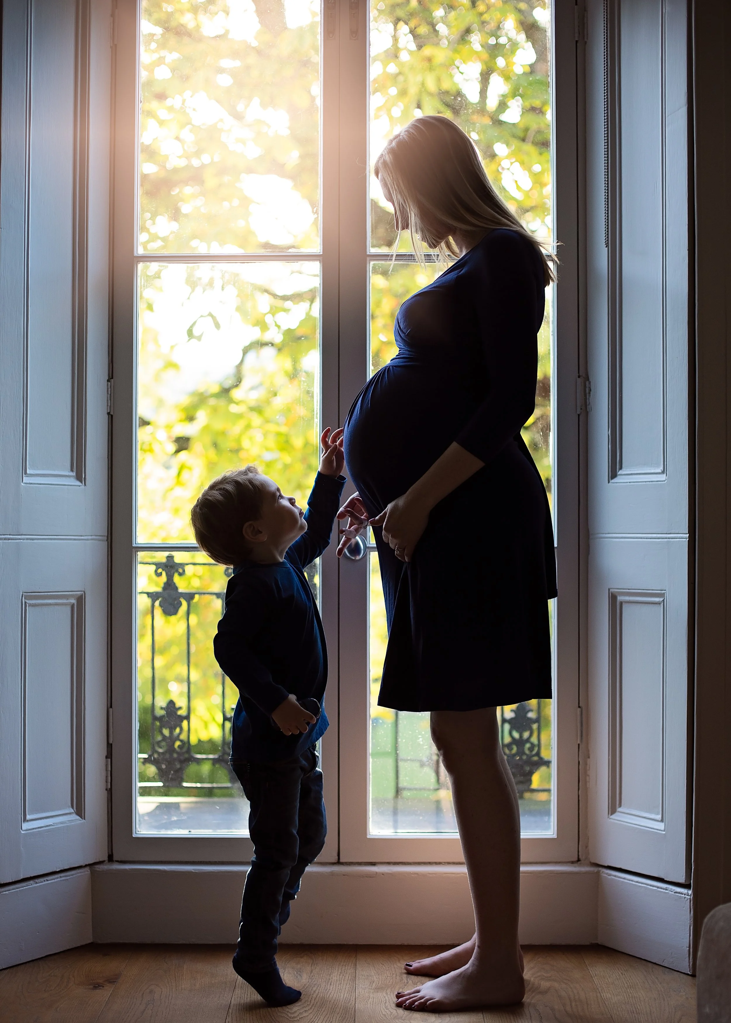 A pregnant woman and a young boy standing by a glass door, looking at each other, with sunlight and greenery visible outside.