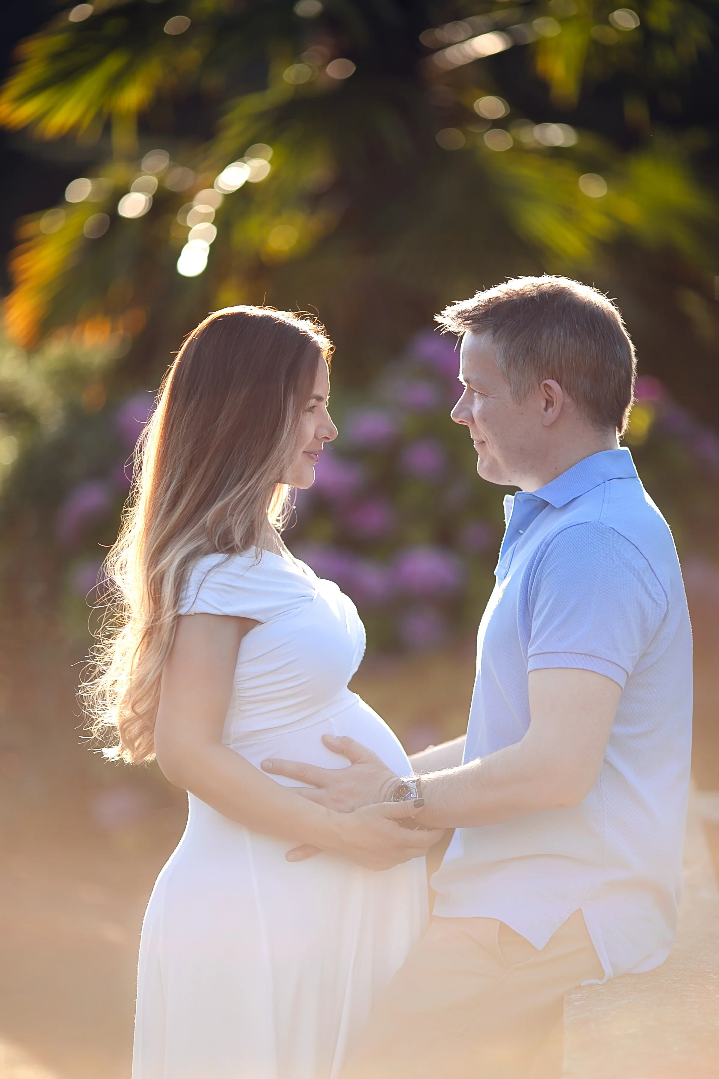 A pregnant woman and a man gazing at each other outdoors, with the woman cradling her belly and the man holding her waist, surrounded by blurred greenery and purple flowers.