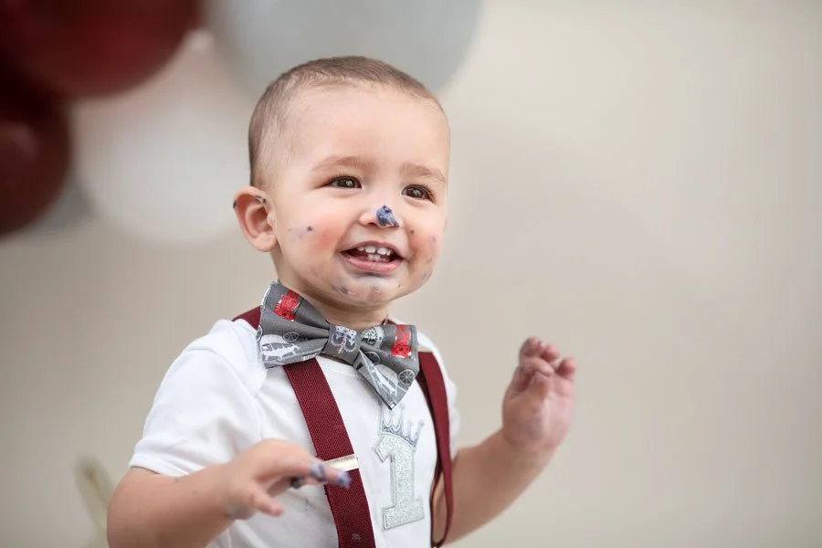 A smiling young boy with short hair, wearing a white shirt and a bow tie, appears to be in a celebratory moment, with paint or food around his mouth and chin, and is holding an object in his hand.
