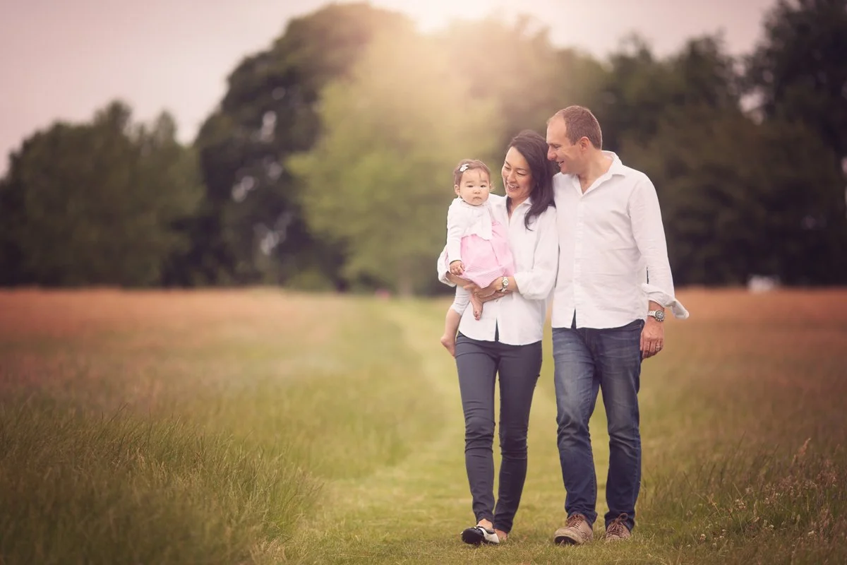 A family of three walking outdoors on a grassy field during sunset, with trees in the background. The mother is holding a young girl, and the father is walking beside them, all smiling.