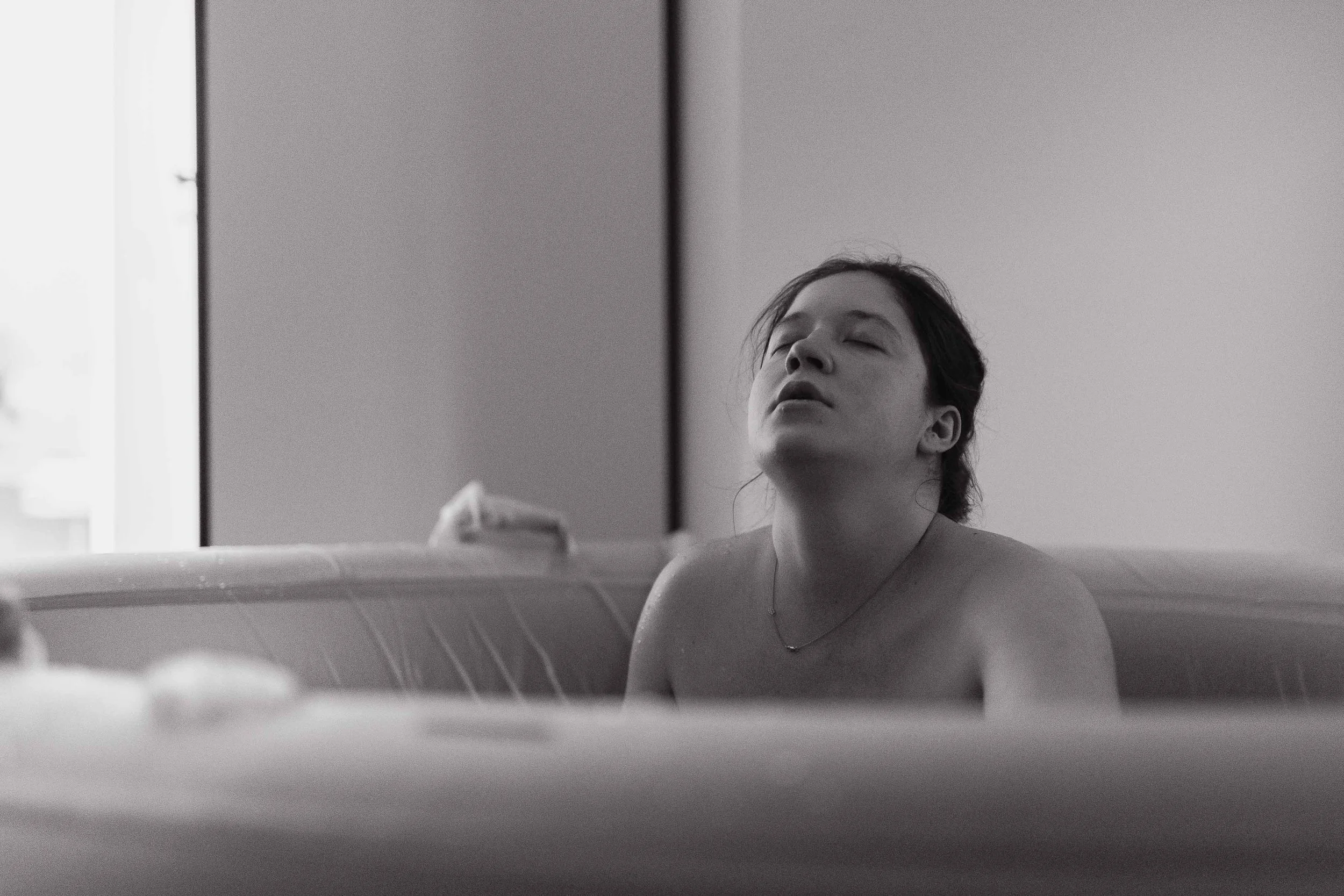 A woman with closed eyes and a relaxed expression, sitting in a bathtub, with part of a mirror visible in the background.