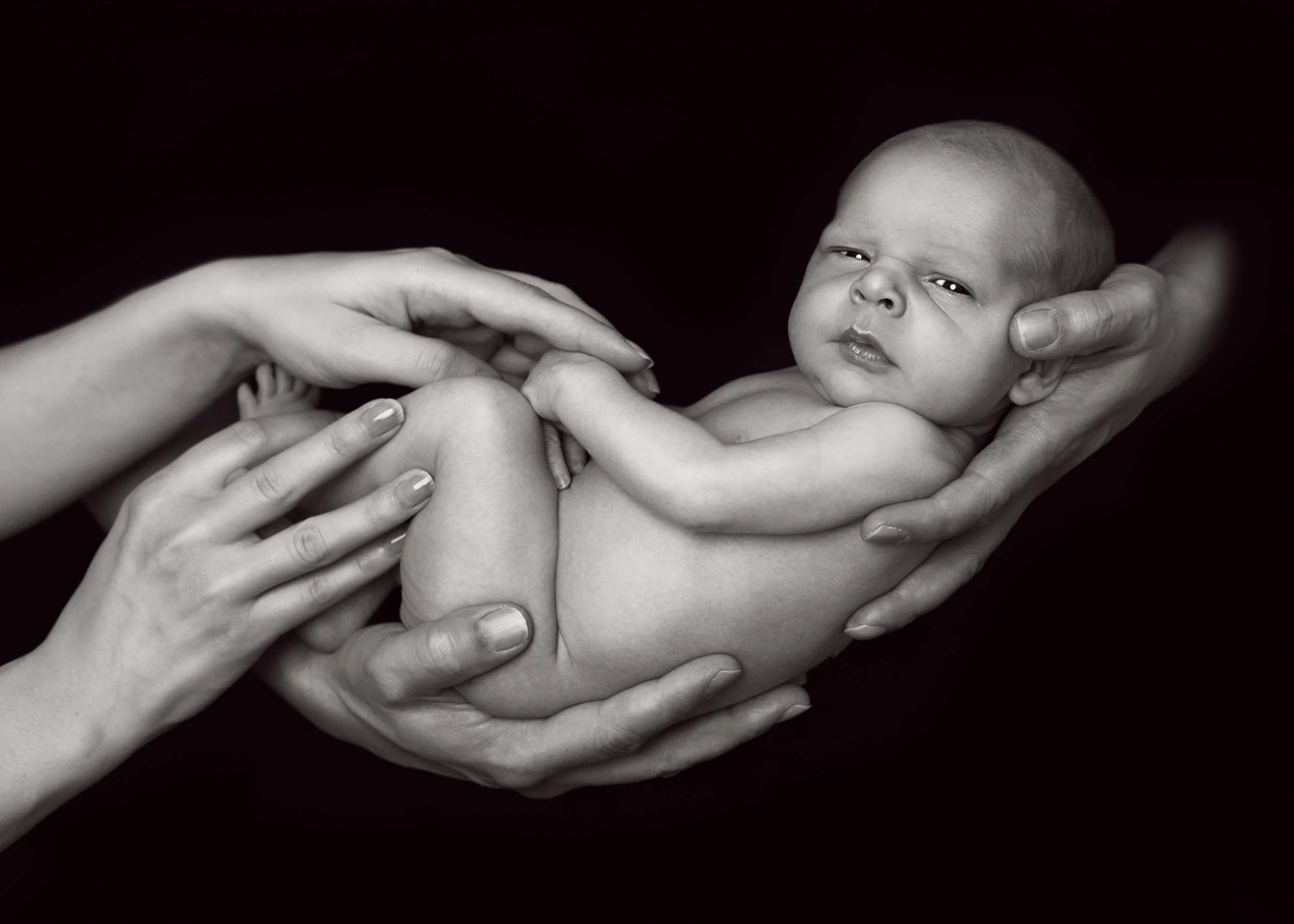 A newborn baby cradled carefully in multiple adult hands against a black background.