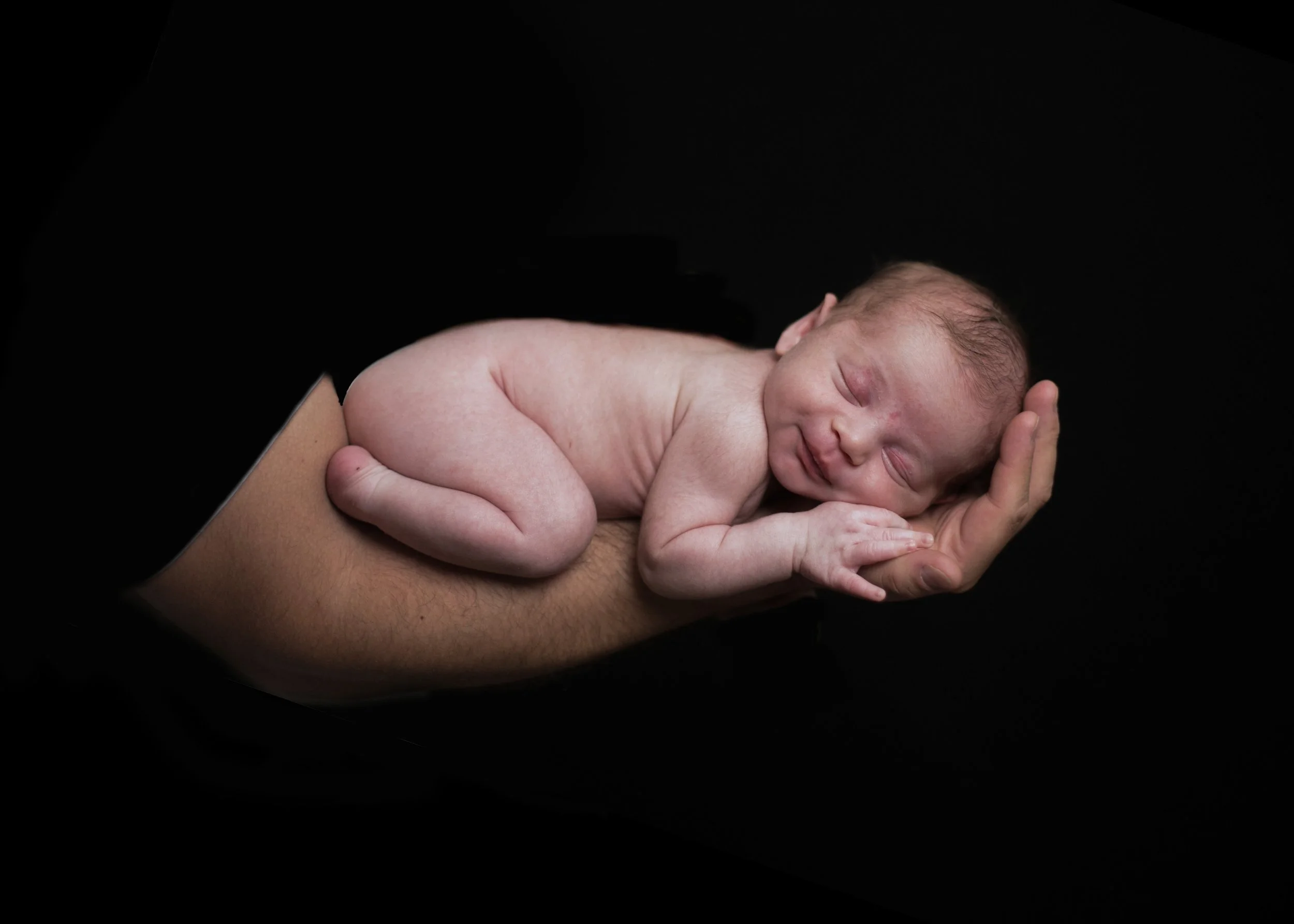 A newborn baby is resting on an adult's arm, with the adult supporting the baby's head and body. The baby appears to be sleeping.