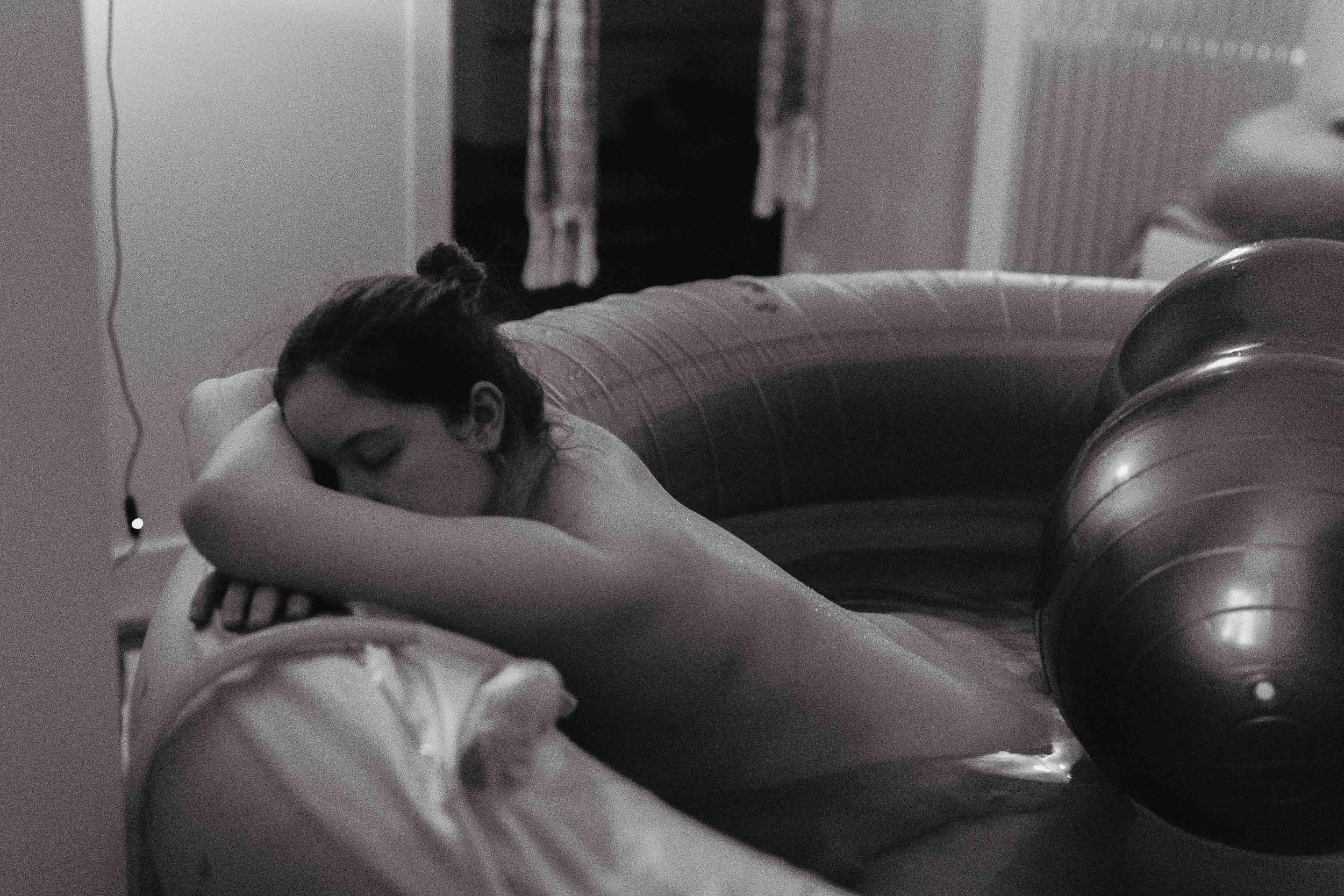 A woman with dark hair pulled back in a bun is resting her head on her crossed arms on a table, with a grainy black-and-white filter effect.