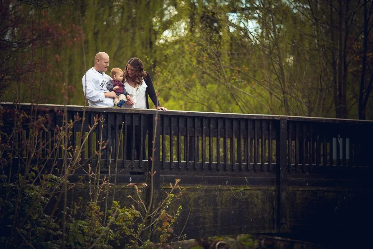 Family of three (father, mother, young child) standing on a wooden bridge in a forested area, enjoying an outdoor moment together.