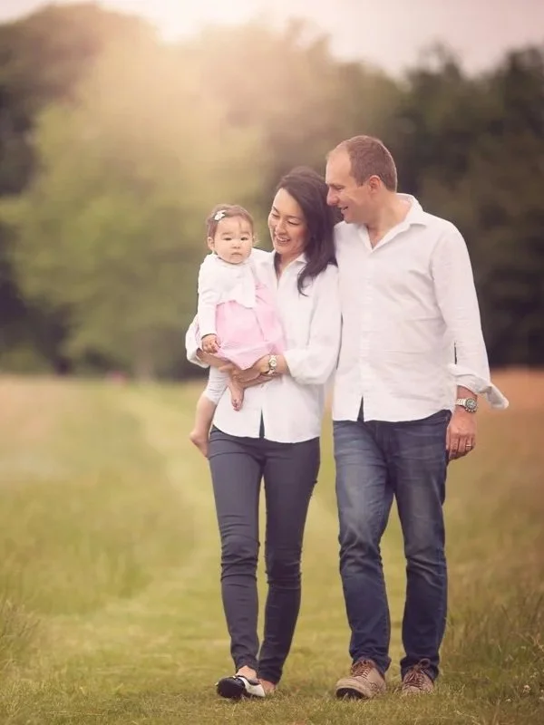 A family of three walking together outdoors, with a man, a woman, and a young girl in the woman’s arms, all smiling.