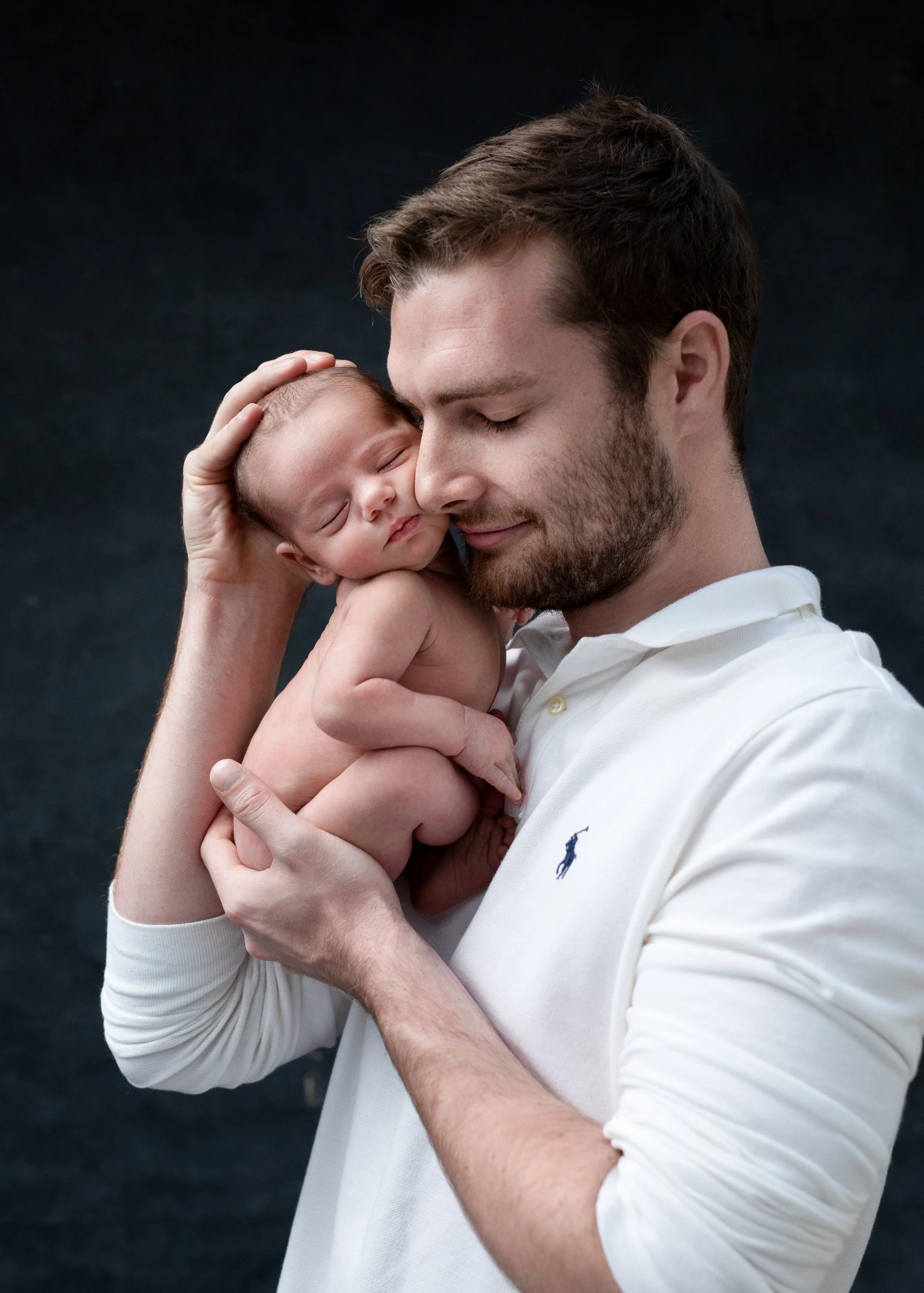 A man with brown hair and a beard holds a newborn baby close to his face against a black background, both with eyes closed and appearing peaceful.