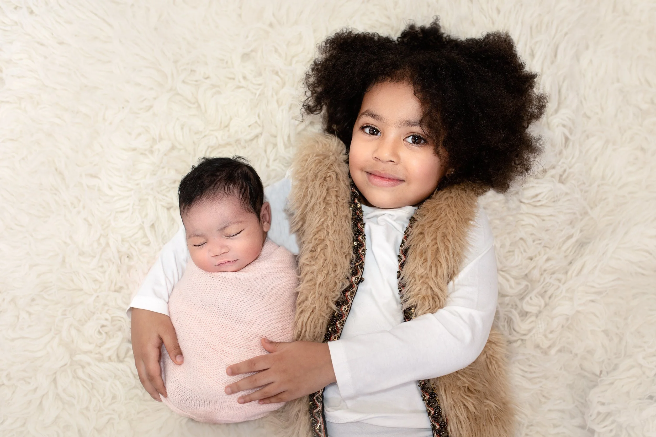 Older sister cuddling her newborn baby sister during an at-home sibling newborn photography session in London by Kate See.