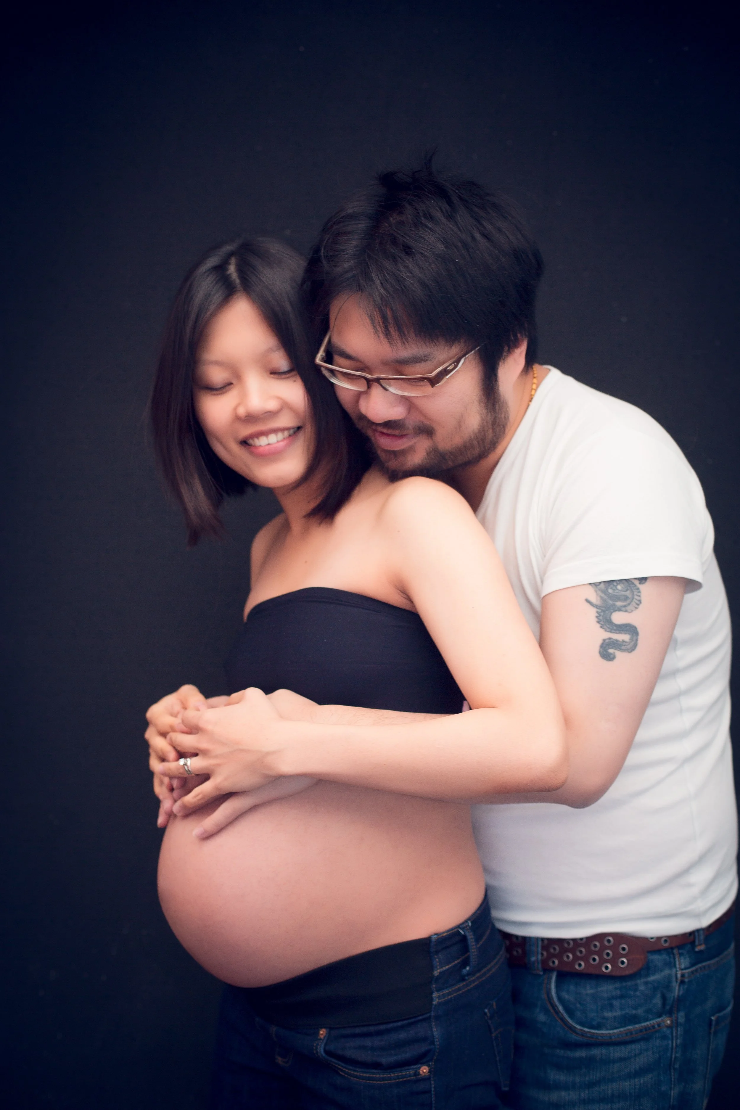 An expecting woman and a man are embracing against a black background. The woman has shoulder-length dark hair and is smiling with her eyes closed. The man, wearing glasses and a white shirt, is resting his forehead on her head, with a tattoo of a dr