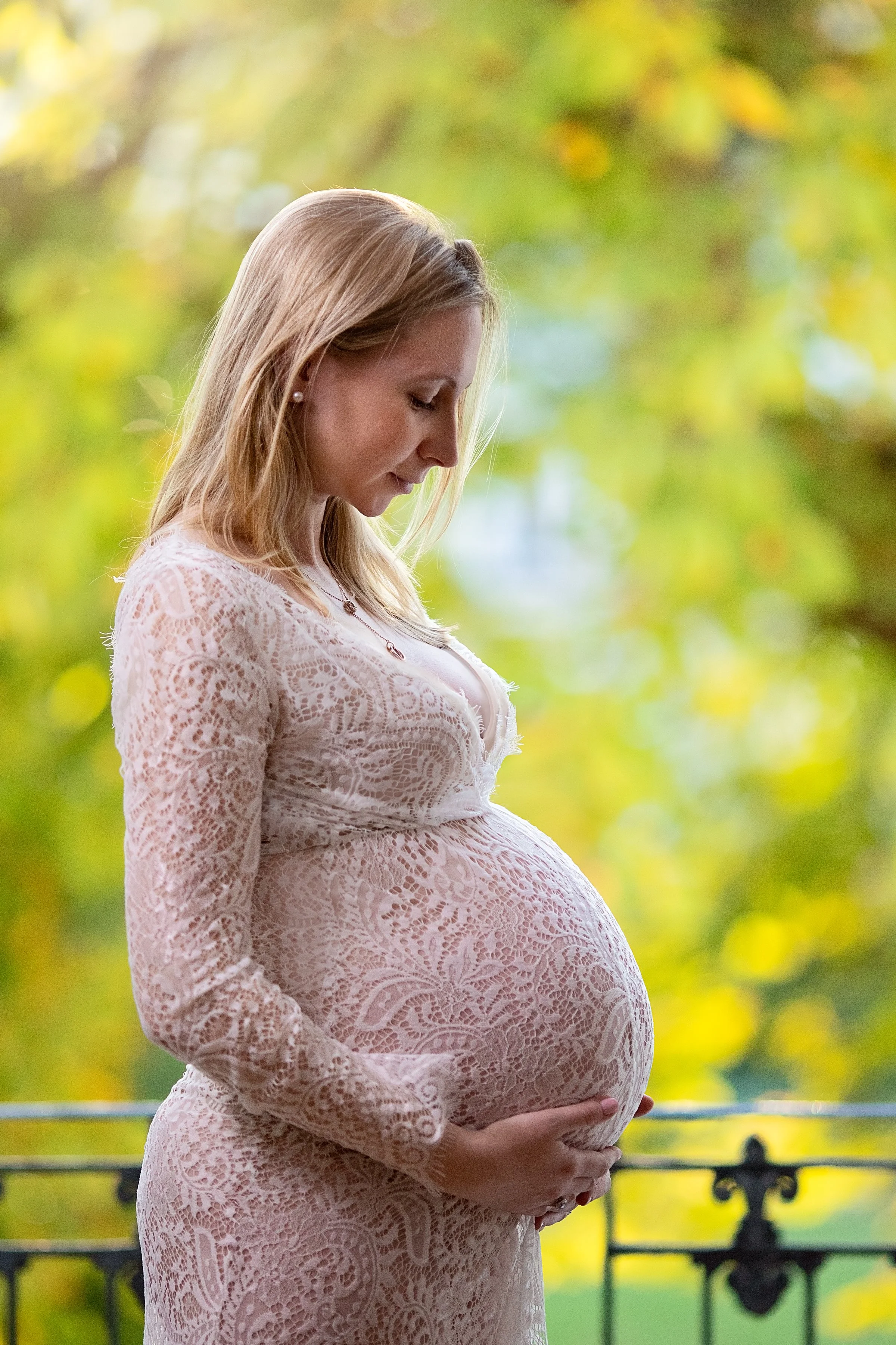 A pregnant woman with light brown hair wearing a lace dress, holding her belly with both hands, standing outdoors in front of a blurred green and yellow foliage background.