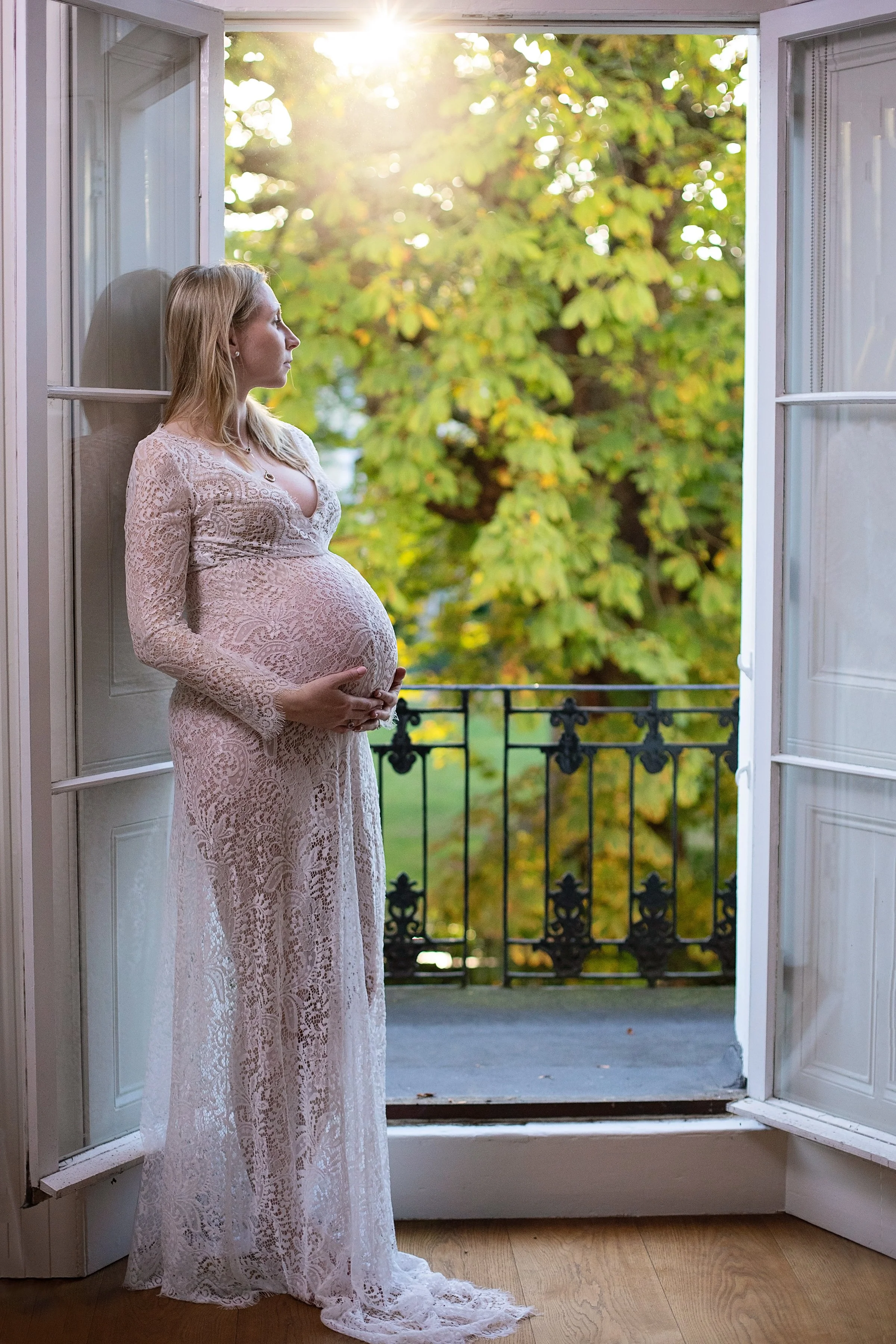 A pregnant woman in a white lace dress stands in front of open French doors, looking outside at a lush green tree and sunlight.
