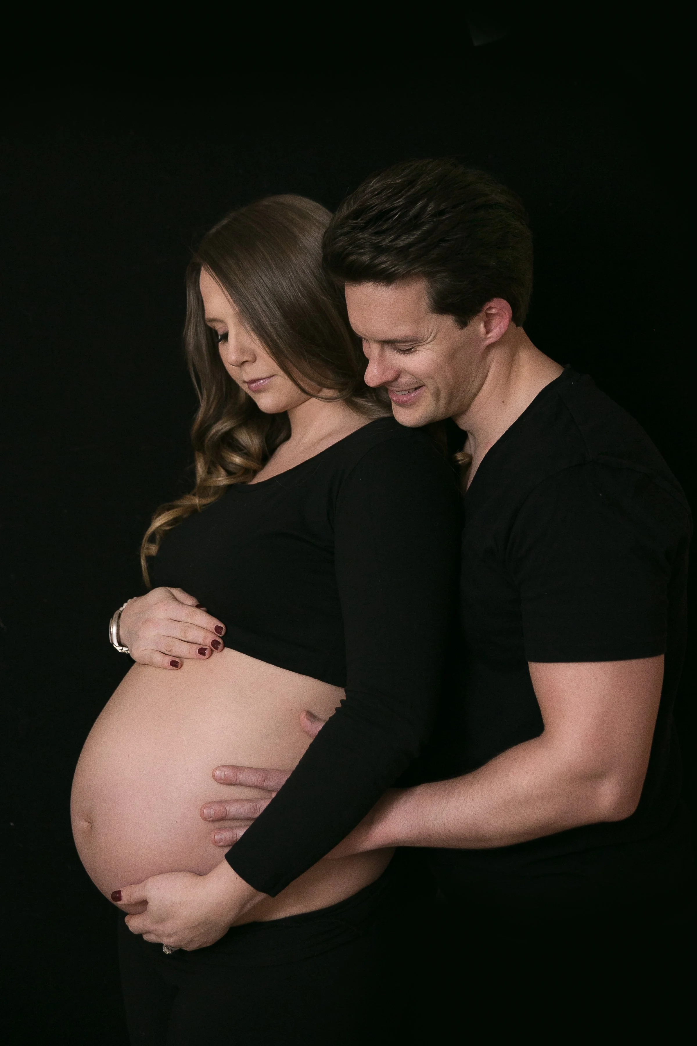 A pregnant woman with long wavy brown hair and a man with short dark hair standing against a black background. The woman is holding her belly and the man is embracing her from behind, both smiling gently with their eyes closed.