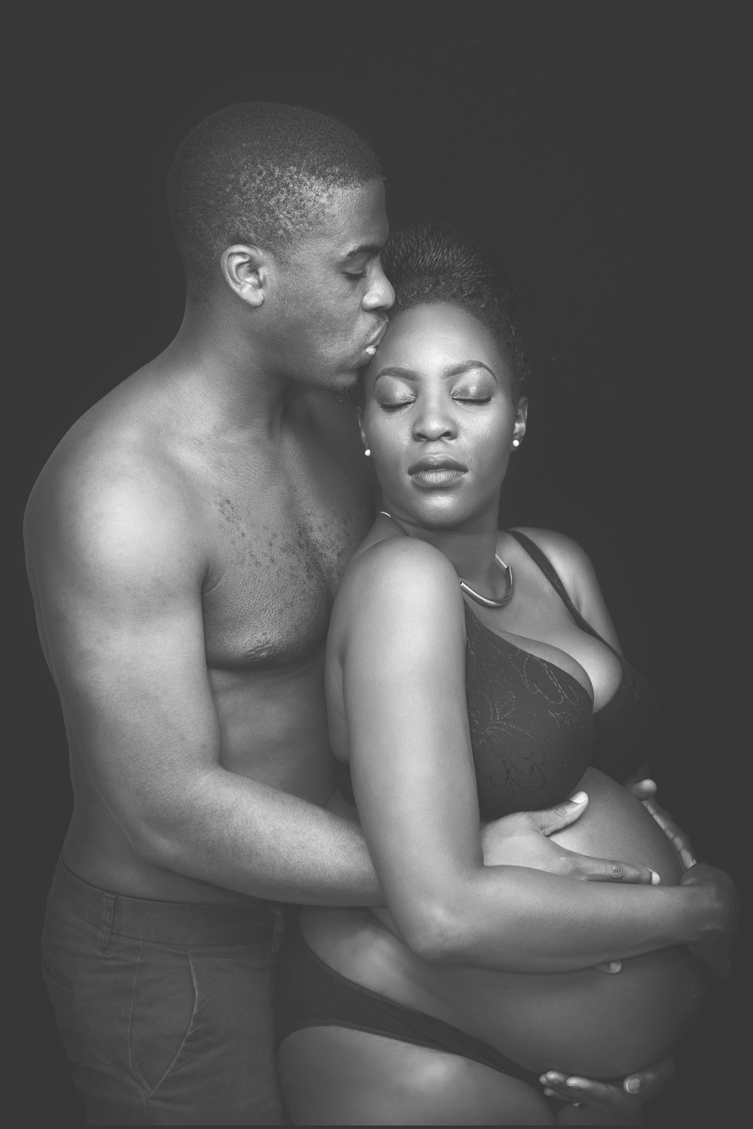 A black and white photograph of a shirtless man with short hair and a woman in a black lace dress, with her eyes closed, both with eyes closed and the man's forehead touching the woman's forehead, against a plain dark background.