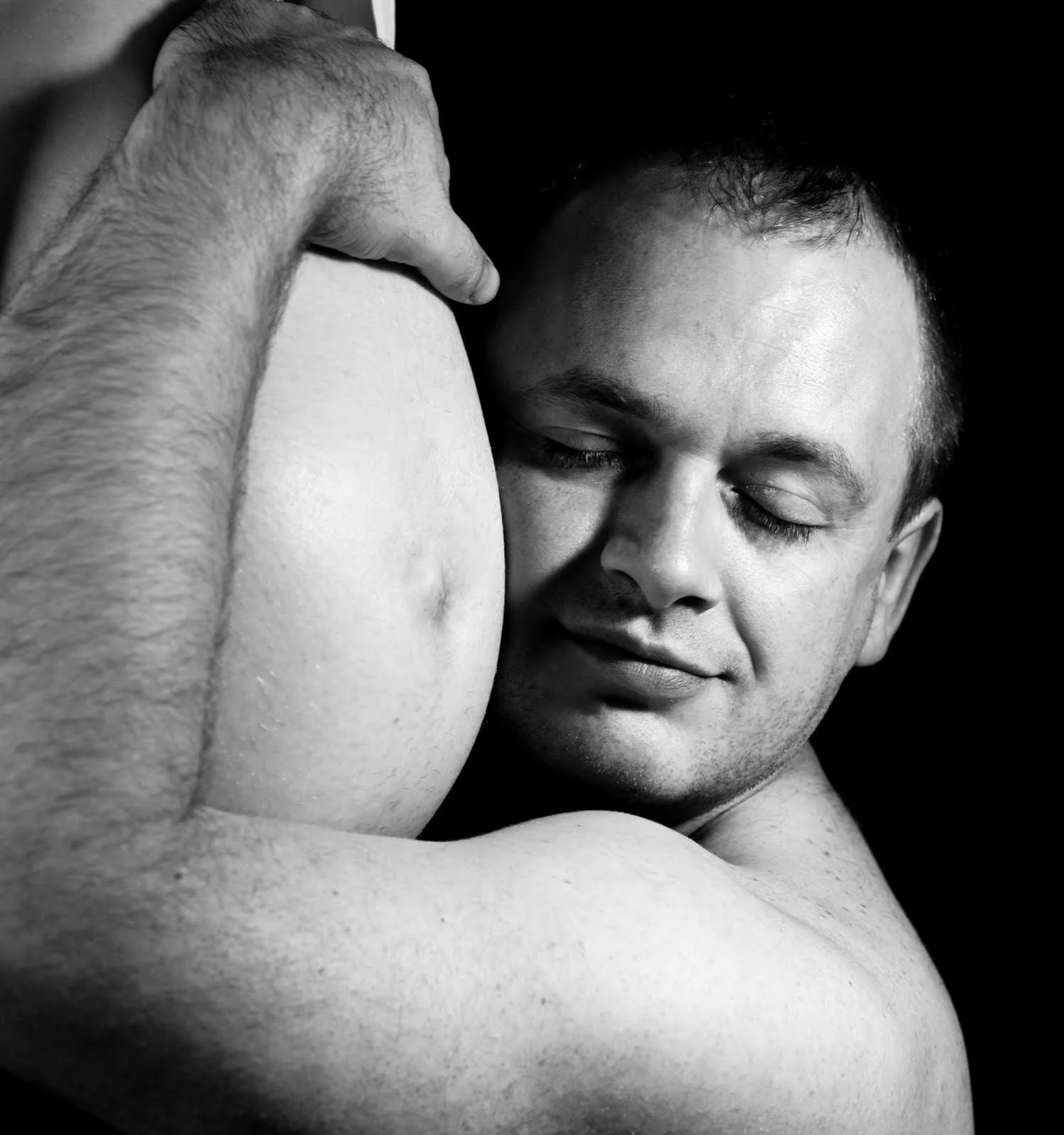 Black and white close-up of two men embracing, with one man’s face resting against the other’s shoulder and a gentle smile on his face.