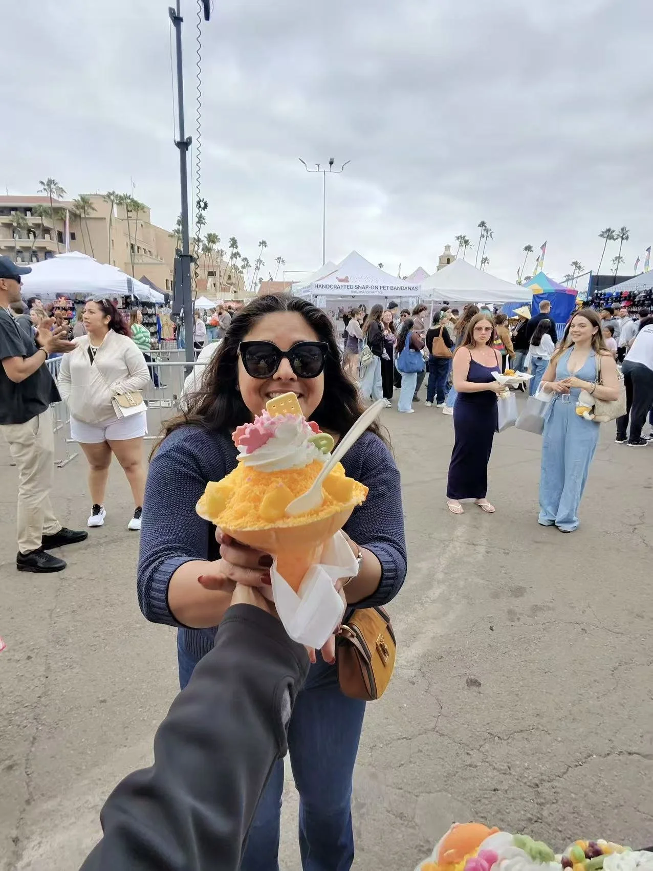 Woman in sunglasses smiling and holding a large shaved ice dessert topped with whipped cream, fruit, and candy at an outdoor fair or festival with many people and vendor tents in the background.