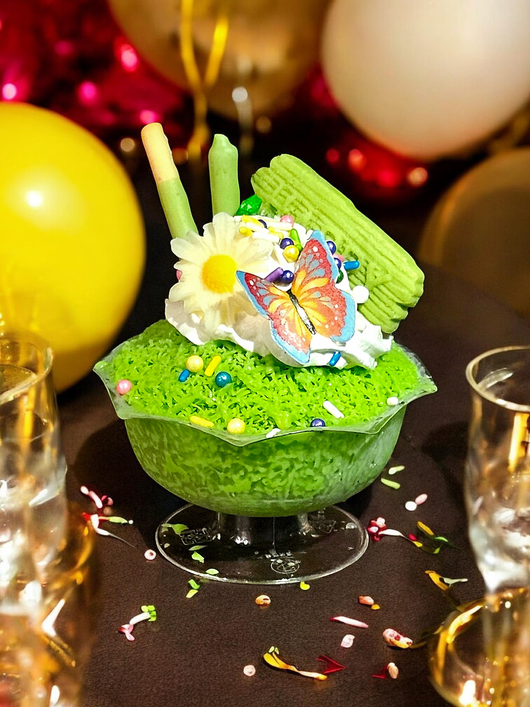 A colorful birthday cake decorated with a butterfly, white flowers, green biscuit sticks, and rainbow sprinkles. The cake is in a green container and surrounded by glittering confetti and two shot glasses with a dark table background.
