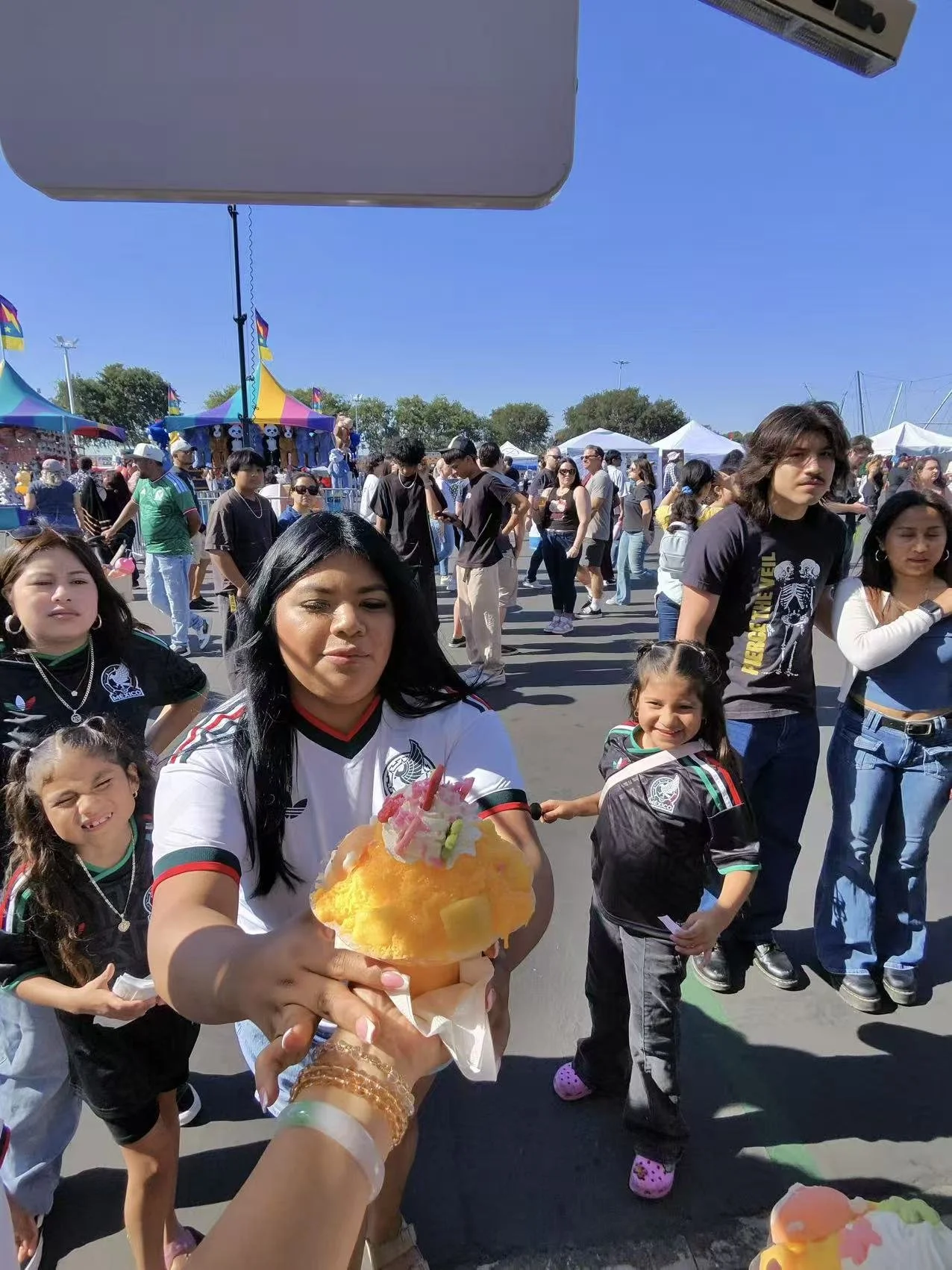 A woman holding a colorful shaved ice dessert at an outdoor festival with a crowd, tents, and carnival booths in the background on a sunny day.