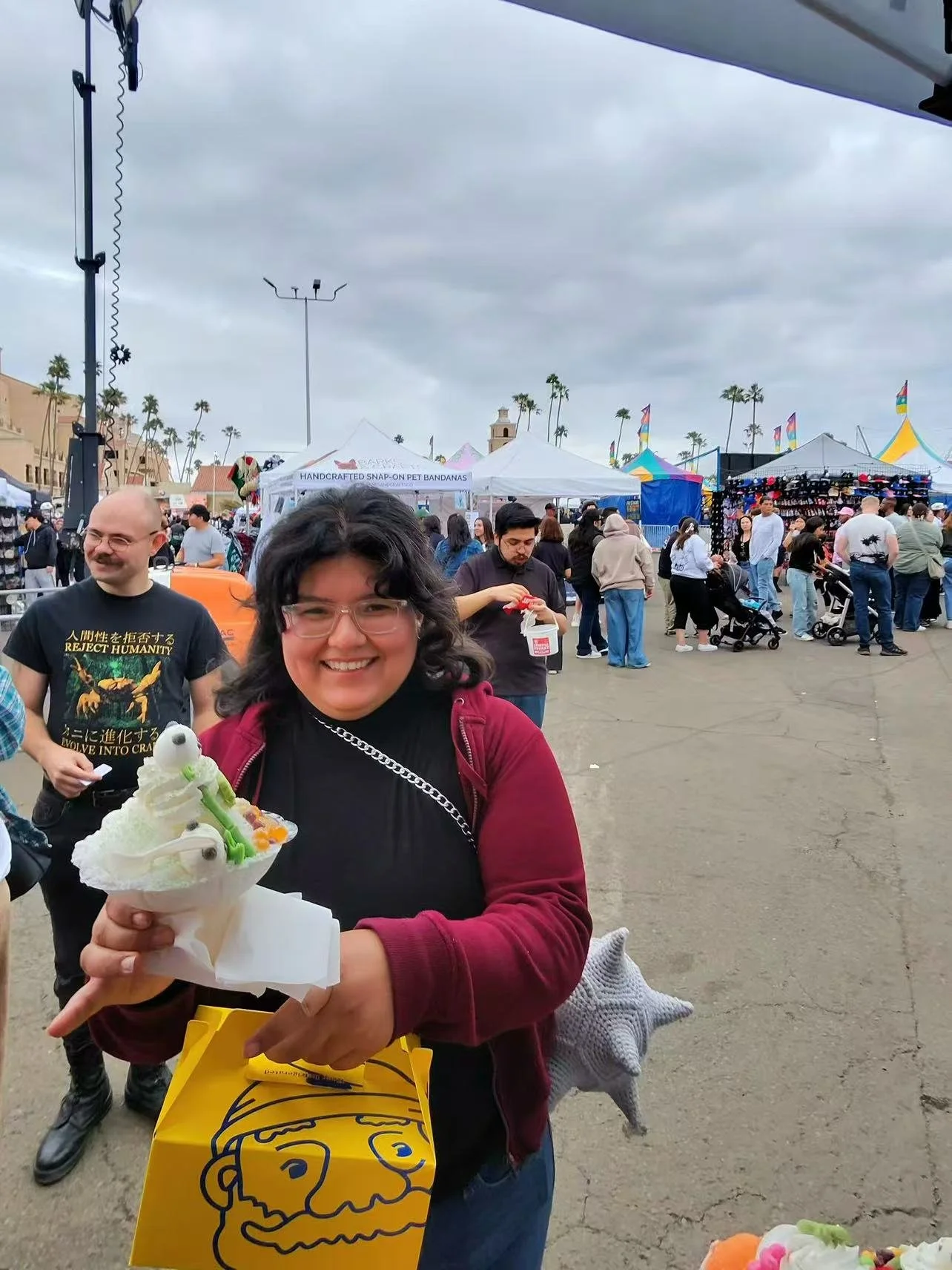 A woman smiling and holding a dessert cone with colorful toppings at an outdoor event or fair. There are tents, booths, and people in the background, with some pushing strollers and browsing. The sky is cloudy.