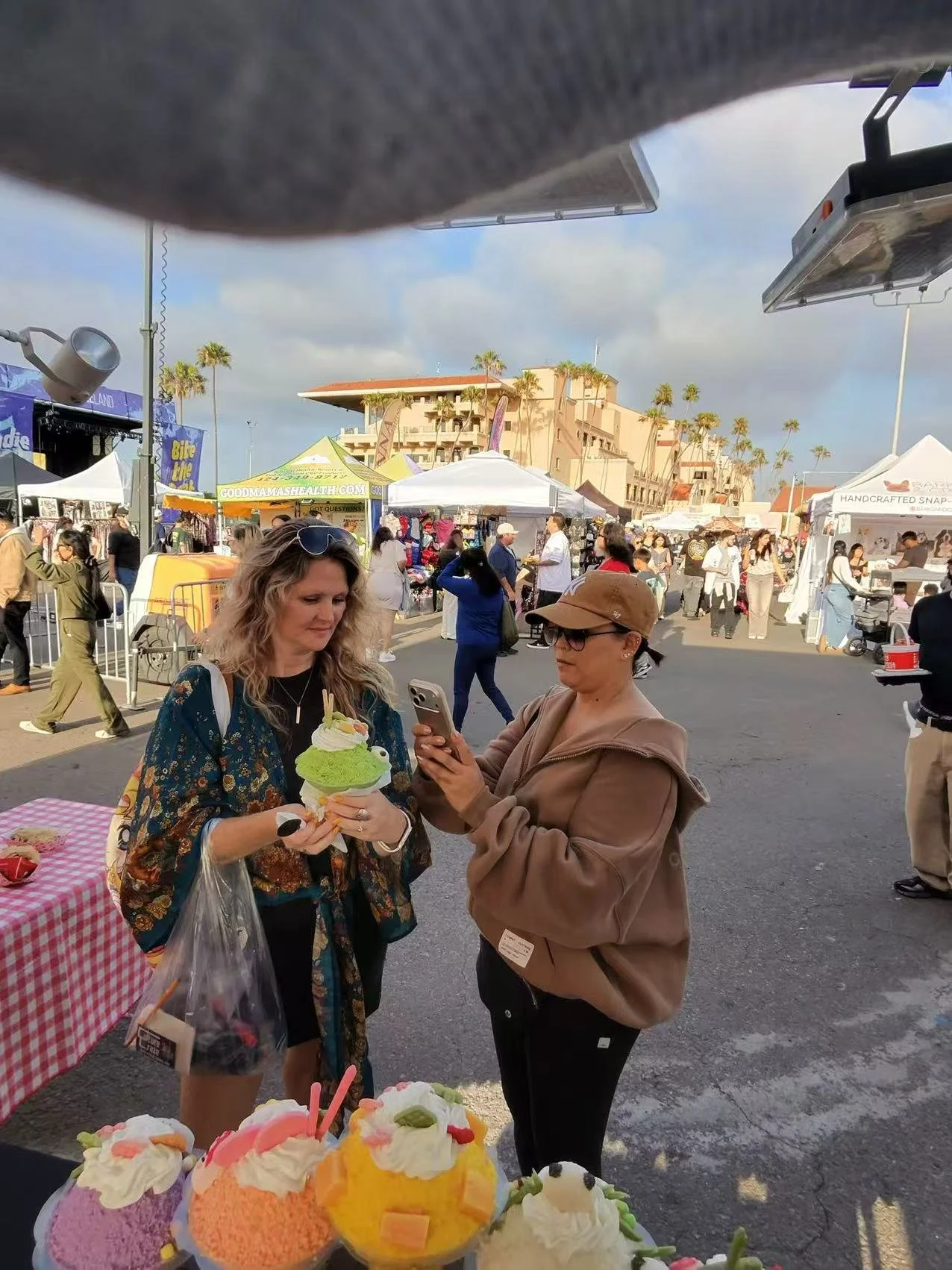 Two women at an outdoor market or festival, with one holding a large colorful ice cream sundae and the other taking a photo of it on her phone. There are various stalls and many people in the background.