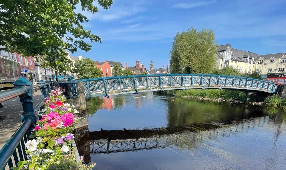 View of Sligo, Ireland, with colourful flowers lining the River Garavogue and a blue pedestrian bridge crossing the water, where the Cairde Sligo Arts Festival and Cairde Visual take place.