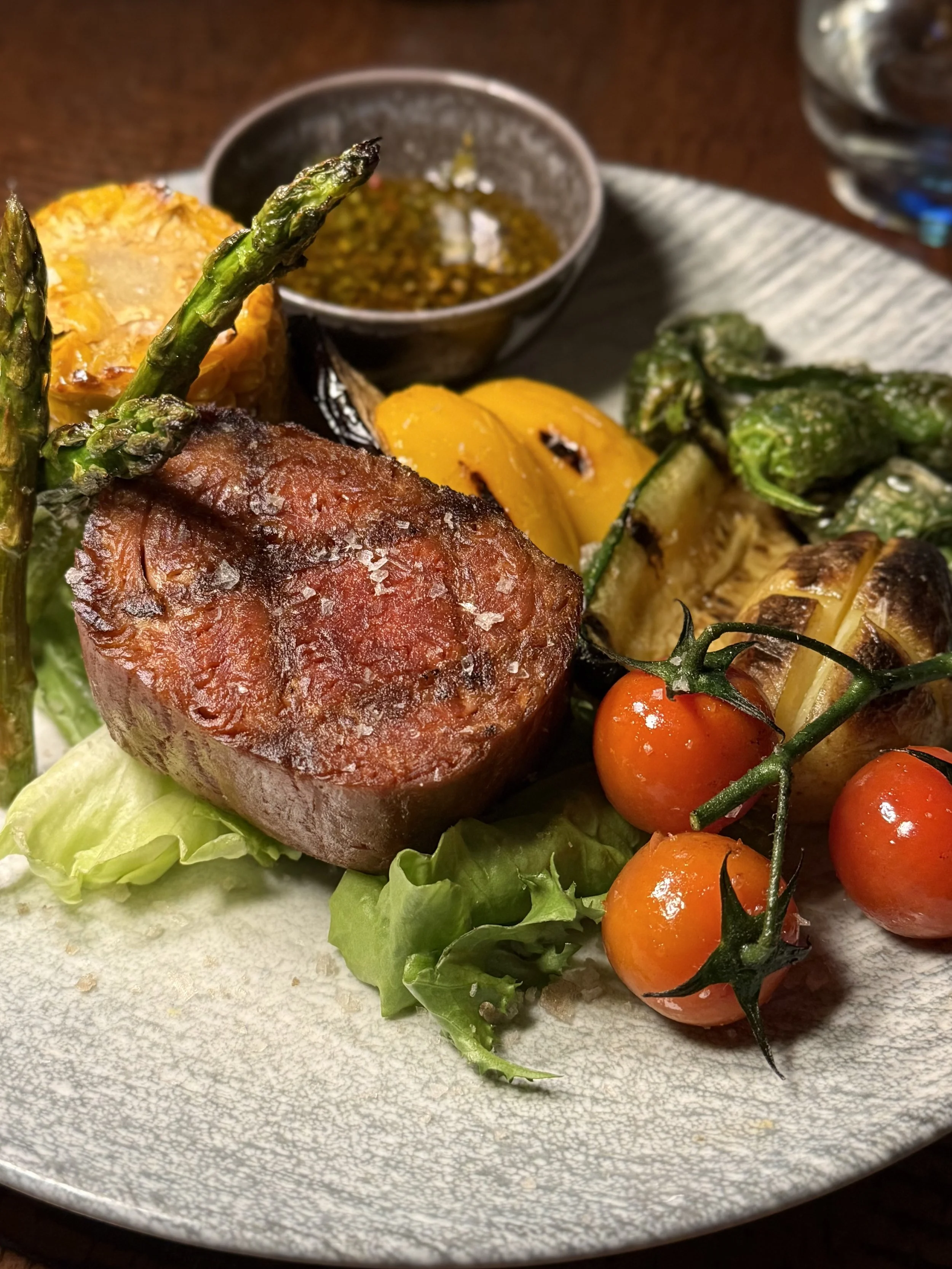 A plate of cooked vegetables, including asparagus, yellow squash, and cherry tomatoes, along with a portion of cooked vegan steak, served with a small bowl of dressing on a plate.