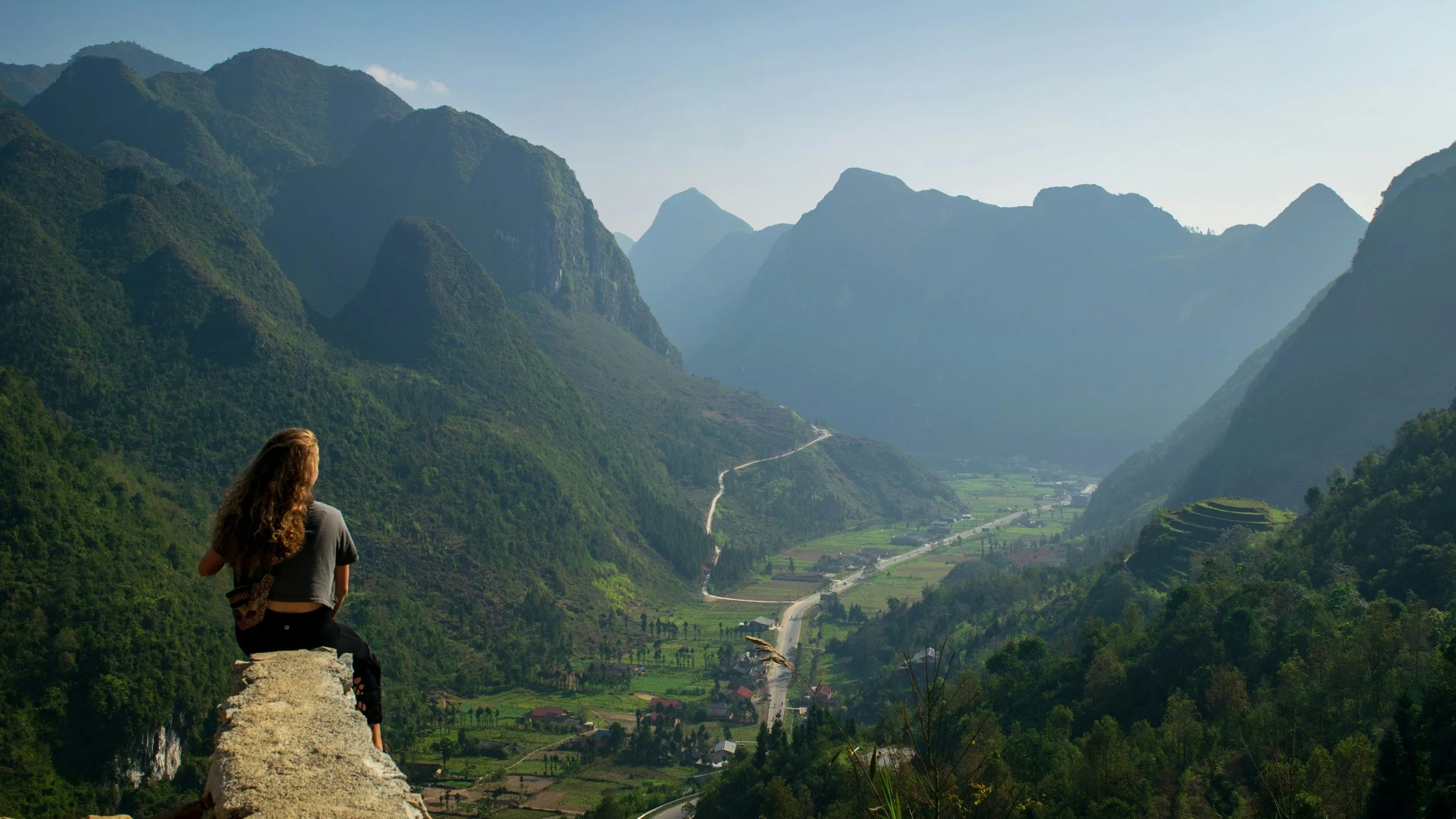 A person with curly hair sitting on a rock, overlooking lush green mountains and a winding road in a valley below.