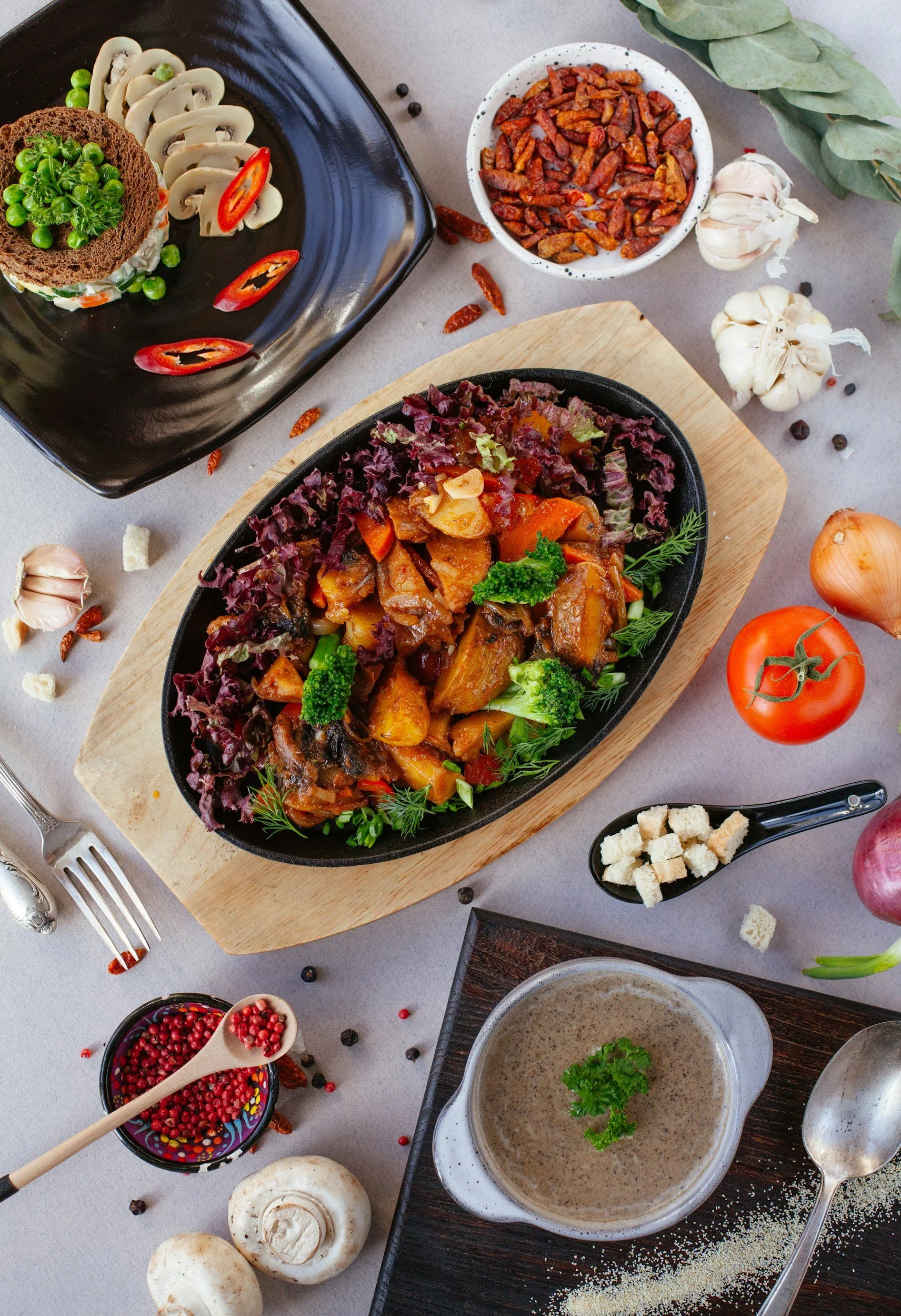 A variety of ingredients and dishes including a vegetable stew, garlic, mushrooms, red onion, berries, and various spices arranged on a light surface.