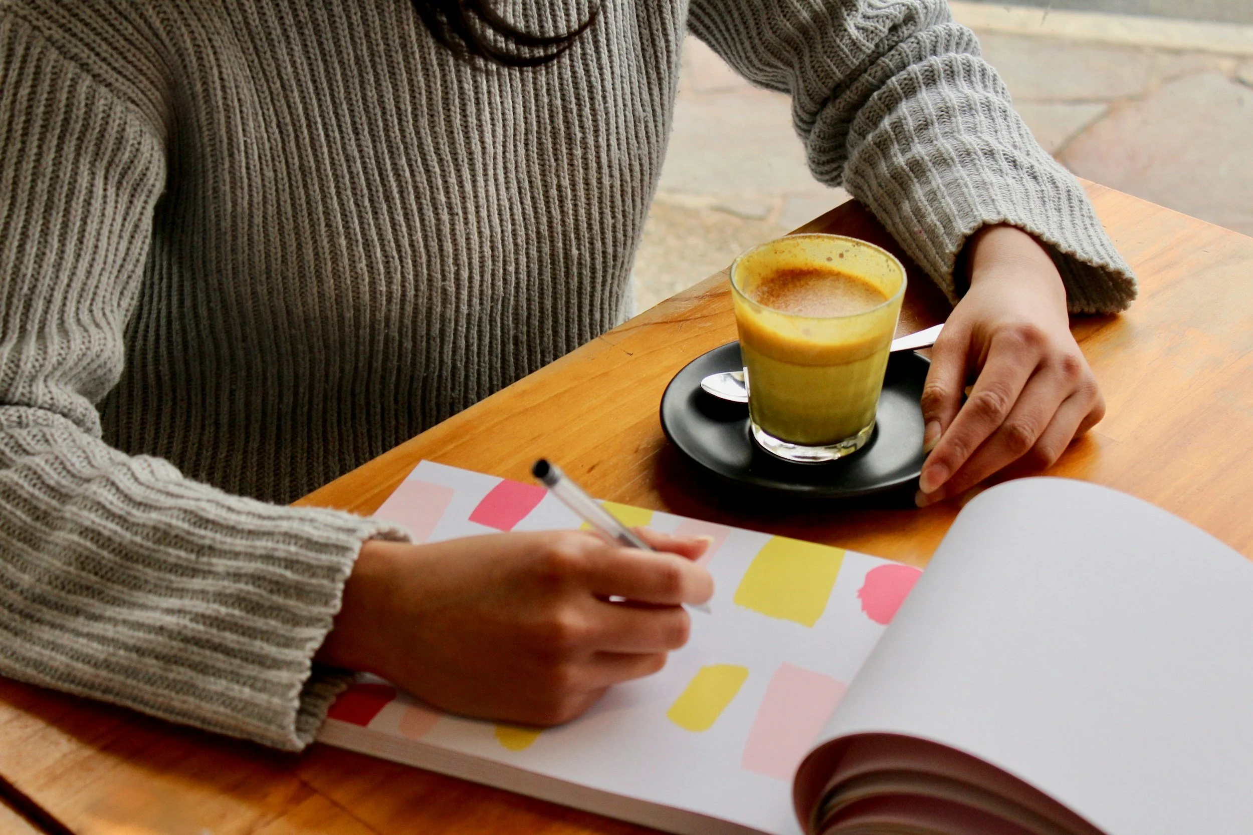 A person wearing a gray, ribbed sweater sitting at a wooden table with a black saucer holding a yellowish drink and silver spoon, writing in a colorful notebook with abstract patterns, while a red coffee cup is partially visible.