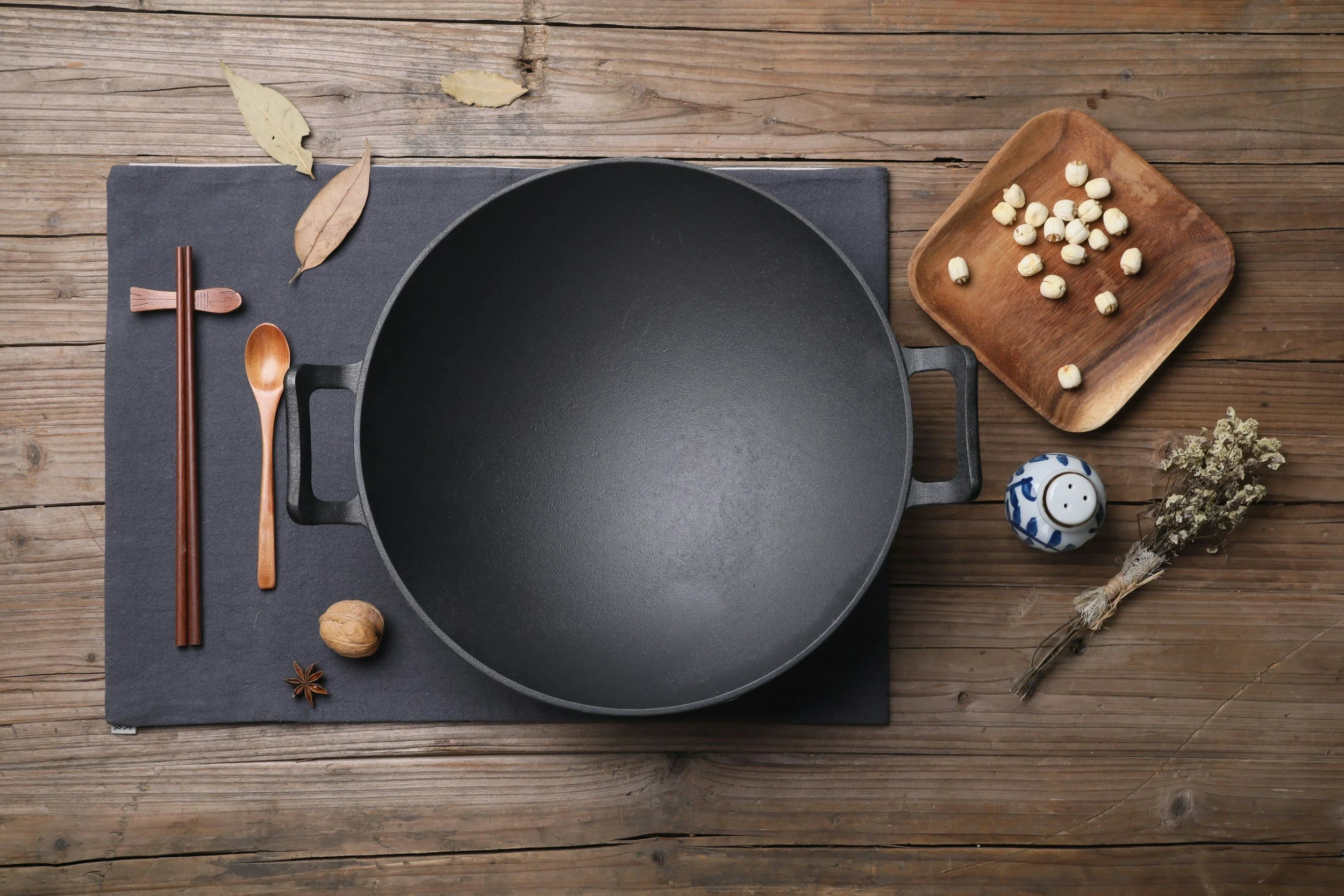 Top-down view of a black cast iron skillet on a black cutting board, with wooden chopsticks, a wooden spoon, dried leaves, a whole nutmeg, a star anise, a small blue and white decorative jar, and a bunch of dried herbs or flowers on a rustic wooden surface.
