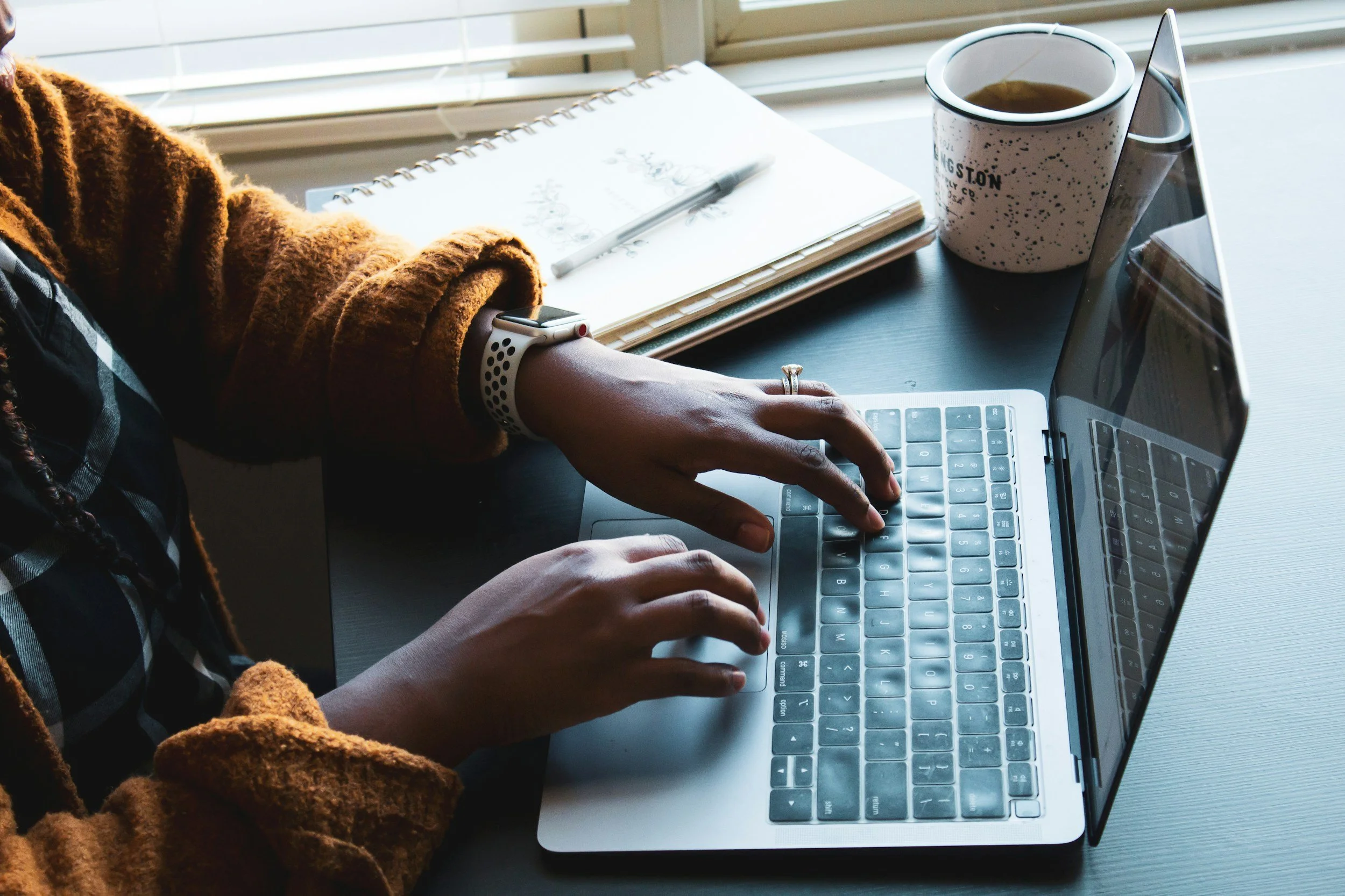 Person working on a silver laptop with a notebook, pen, and coffee mug on the desk.
