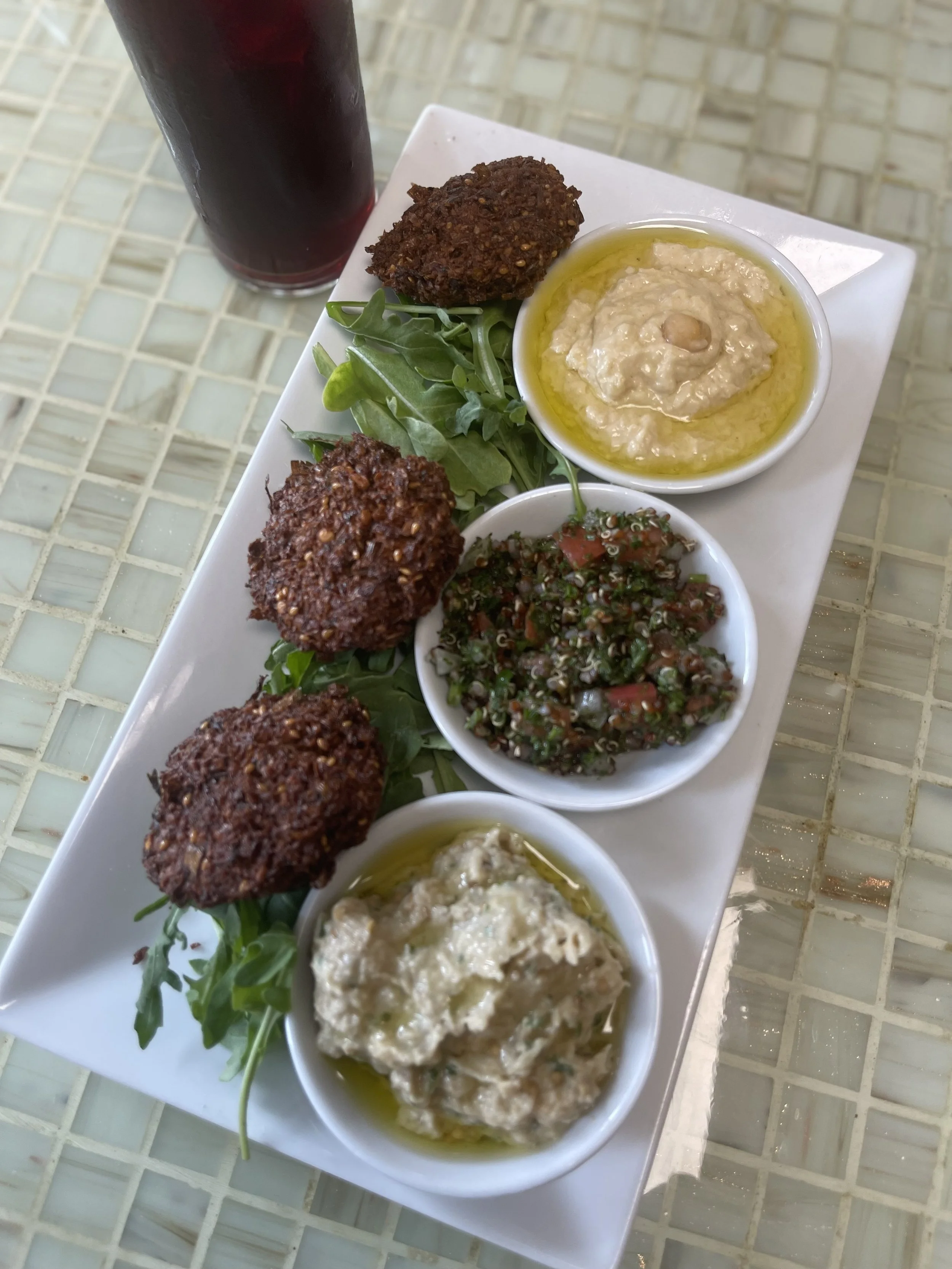 A rectangular white plate with four small bowls of different dips, topped with green arugula leaves. The dips include a creamy white one with herbs, a brown one with grains, a red chopped vegetable salad, and a beige one in the foreground. There is a tall glass of dark red beverage in the background.