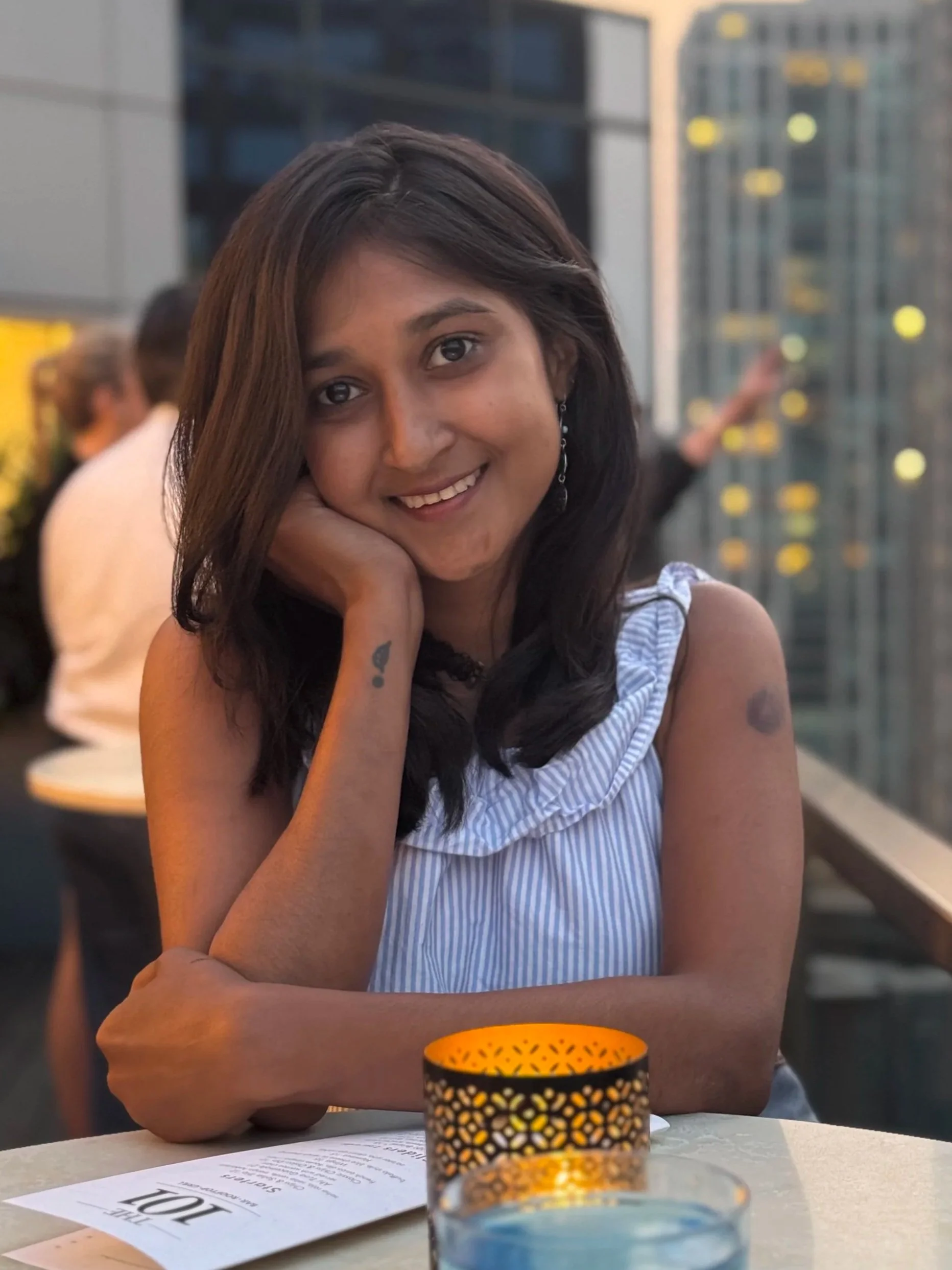 A young woman with dark hair and a tattoo on her wrist, smiling and resting her head on her hand, sitting at a table with a candle holder and a menu, in a social setting with blurred background.