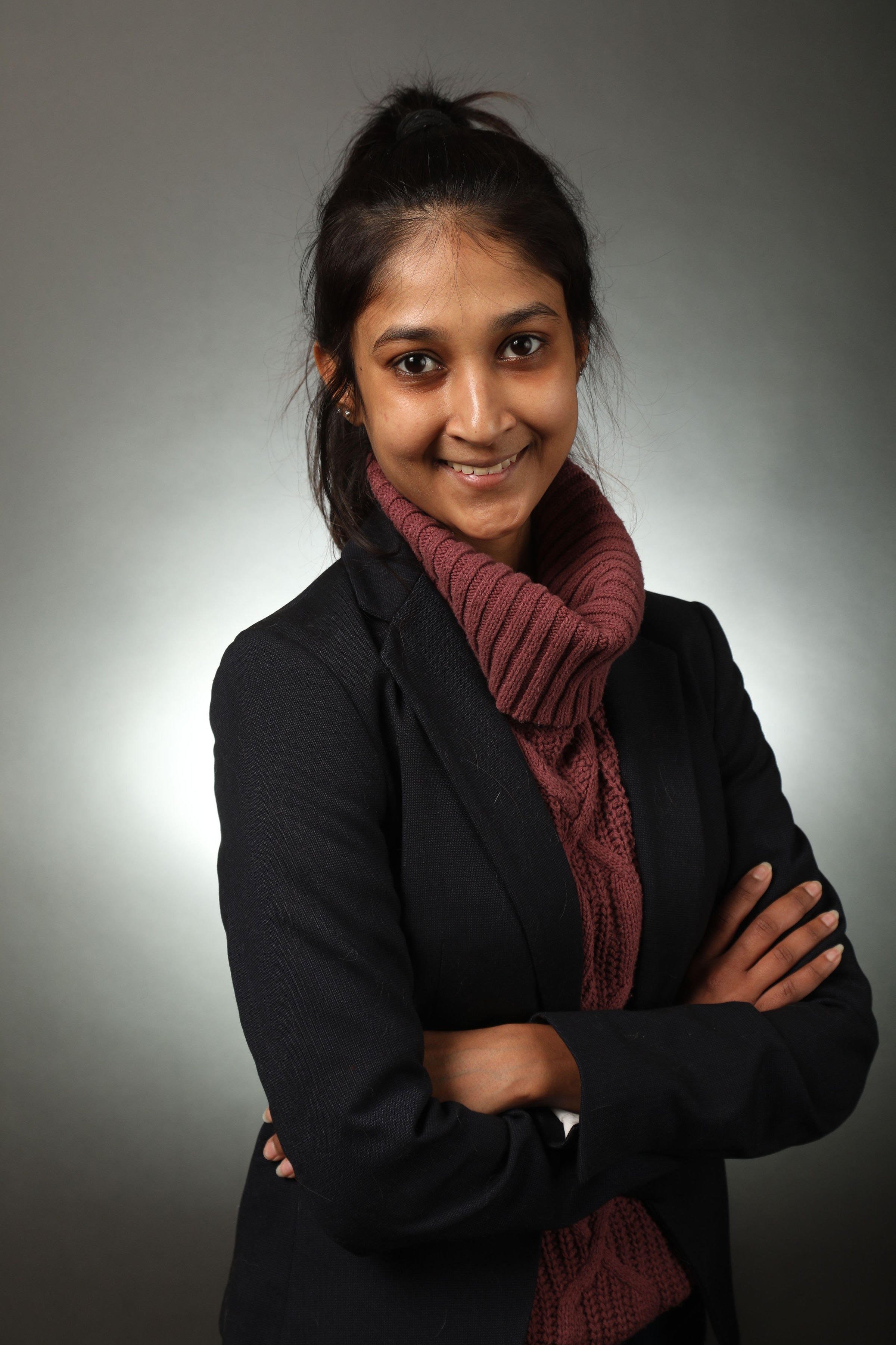 A young woman with dark hair pulled back, smiling, wearing a black blazer and a maroon knit scarf, standing against a plain gray background.