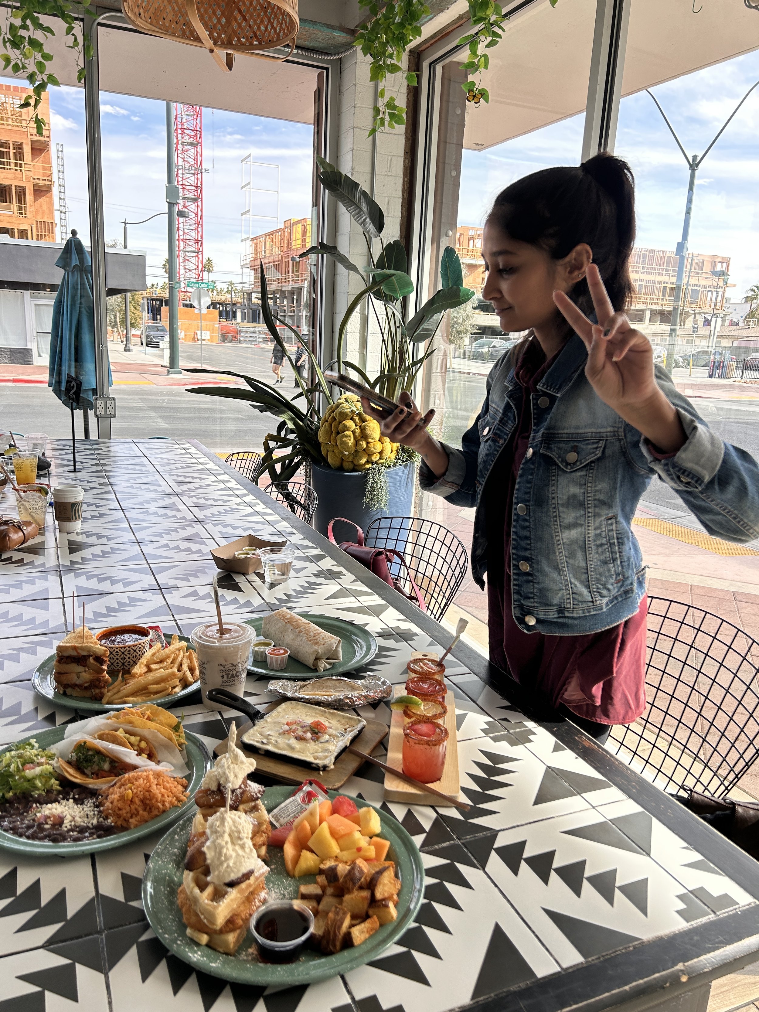 A woman with dark hair tied in a ponytail, wearing a denim jacket, making a peace sign with her right hand and looking at her phone, standing in front of a table filled with various dishes, including tacos, fries, desserts, and drinks, inside a restaurant with large windows and a view of a construction site outside.