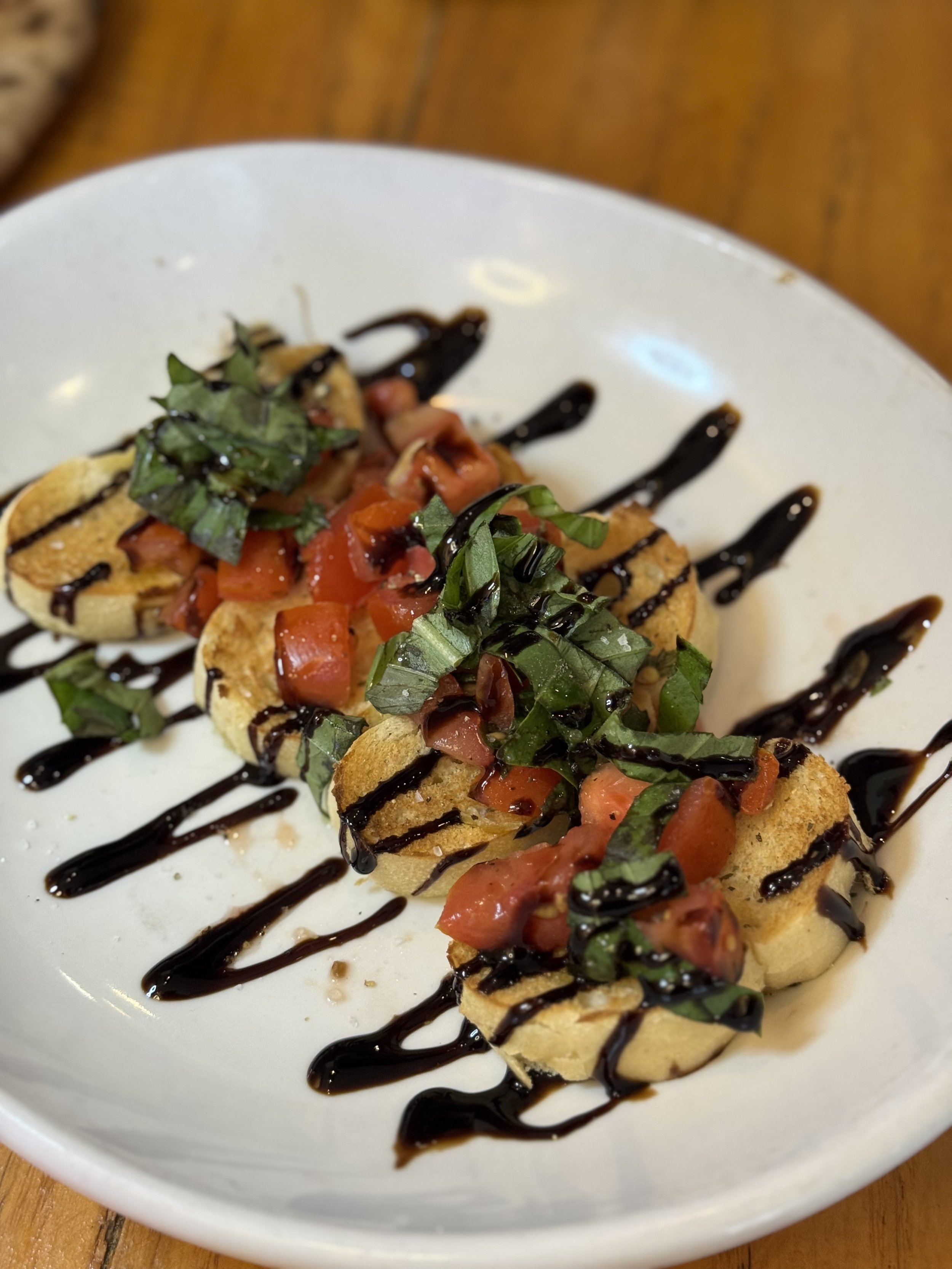 Close-up of a white bowl with toasted baguette slices topped with chopped tomatoes, fresh herbs, and drizzled with balsamic glaze, set on a wooden table.