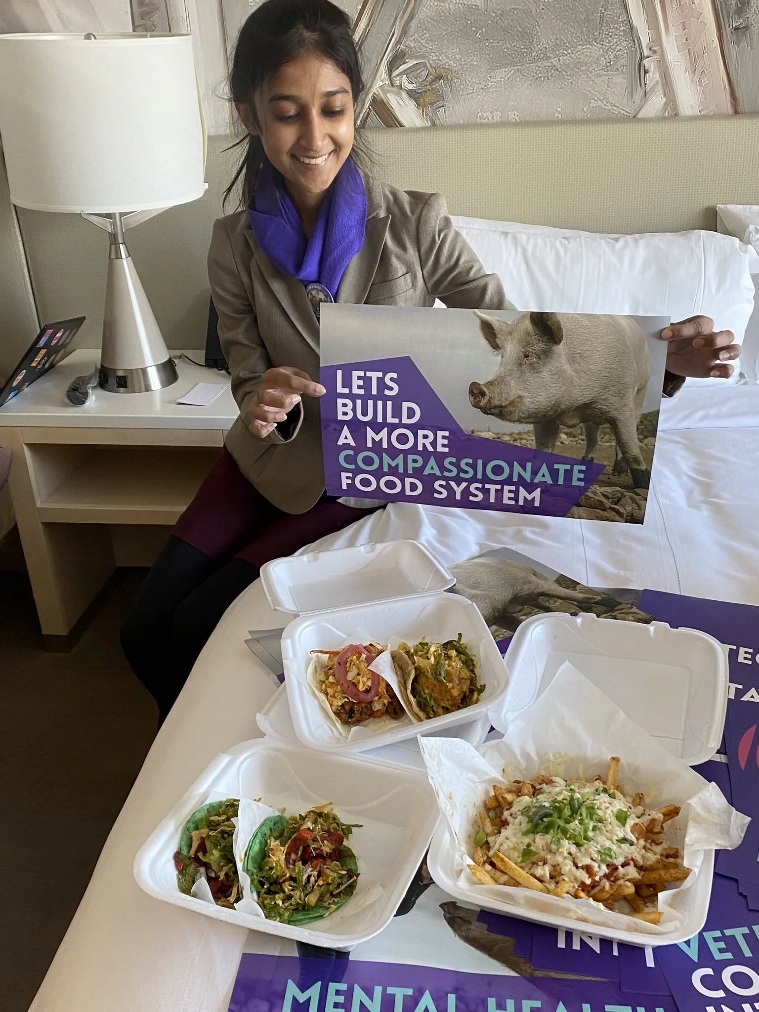 A woman sitting on a bed, holding a sign that says 'Let's build a more compassionate food system' with an image of a pig. On the table in front of her are several food containers with different dishes and salads.