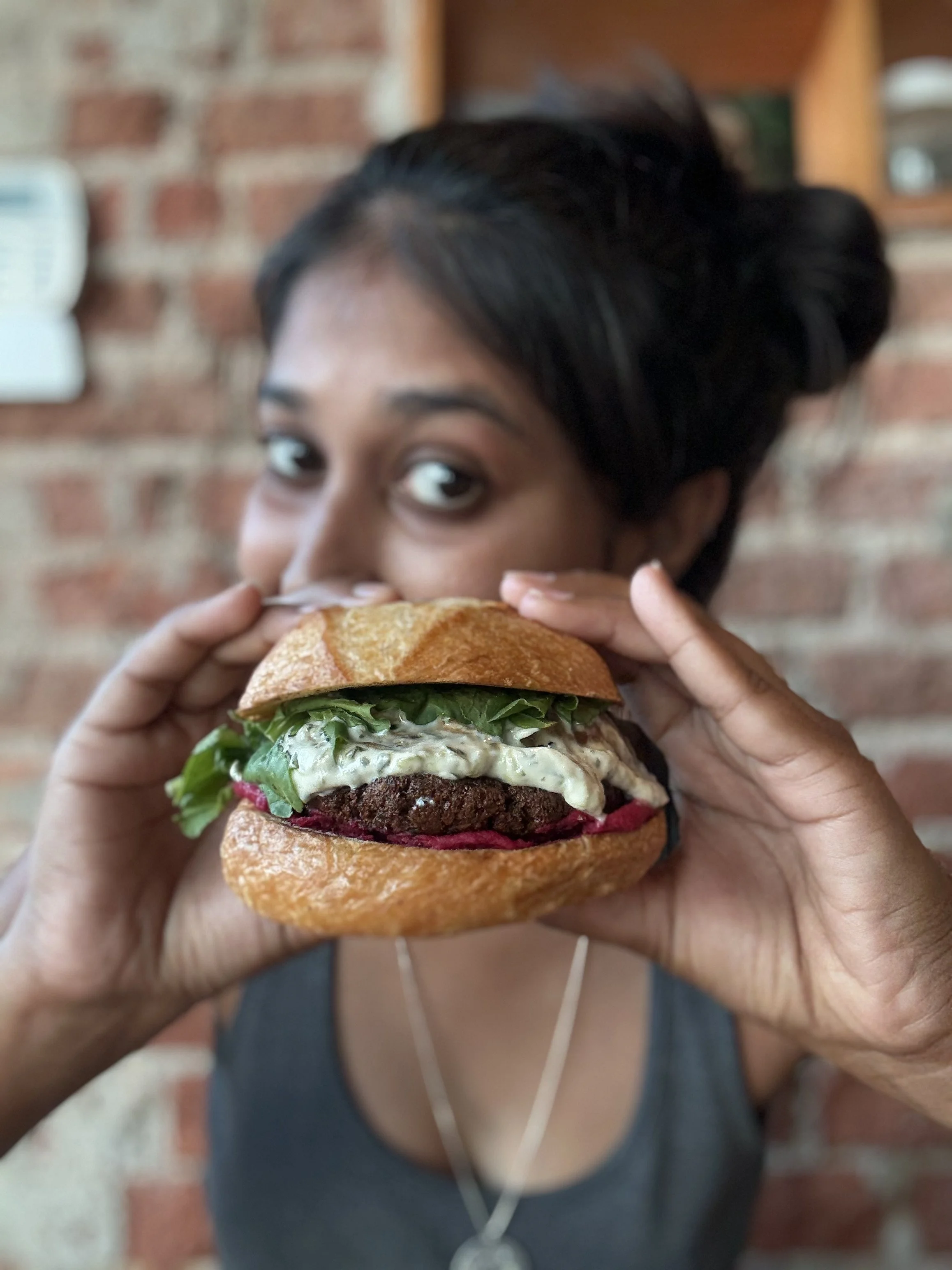 A person with short dark hair holding a large sandwich with lettuce, tomato, cheese, and a thick patty, in front of a brick wall, wearing dark clothing and earphones.