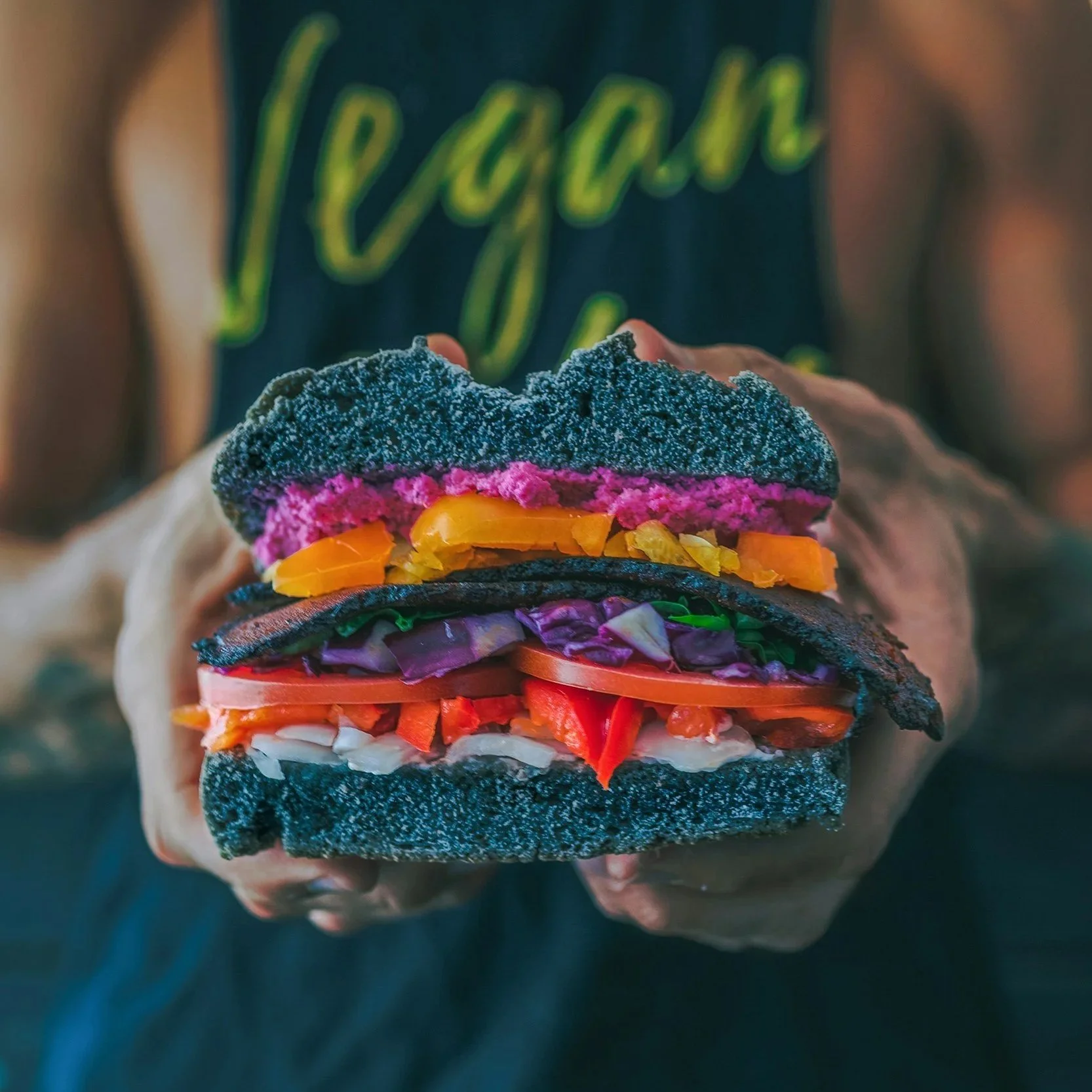 Close-up of a person holding a large colorful burger with black charcoal buns, layers of shredded vegetables, and slices of tomato.