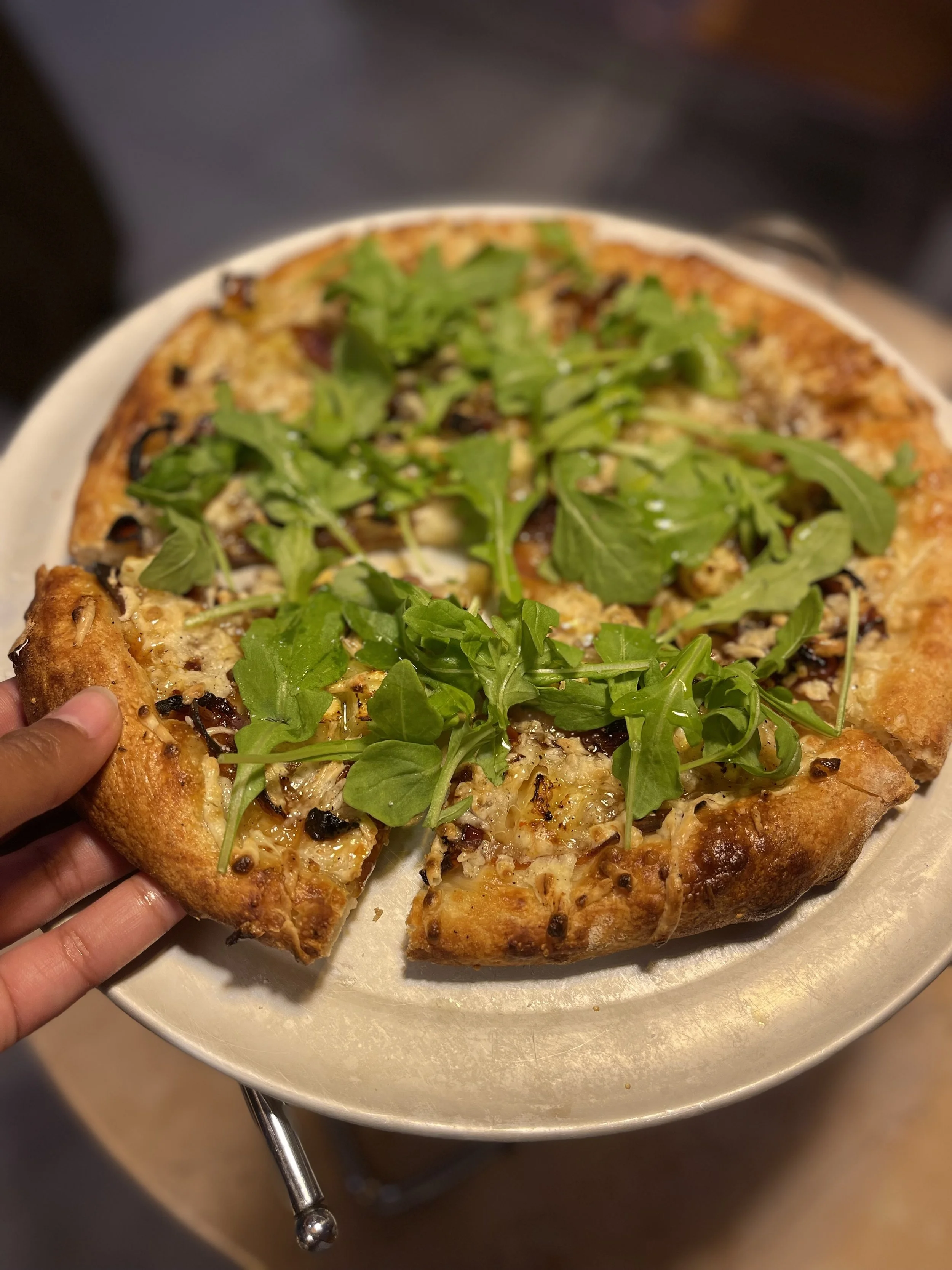 Close-up of a pizza with green arugula on top, placed on a white plate. A person is holding a slice with their fingers.
