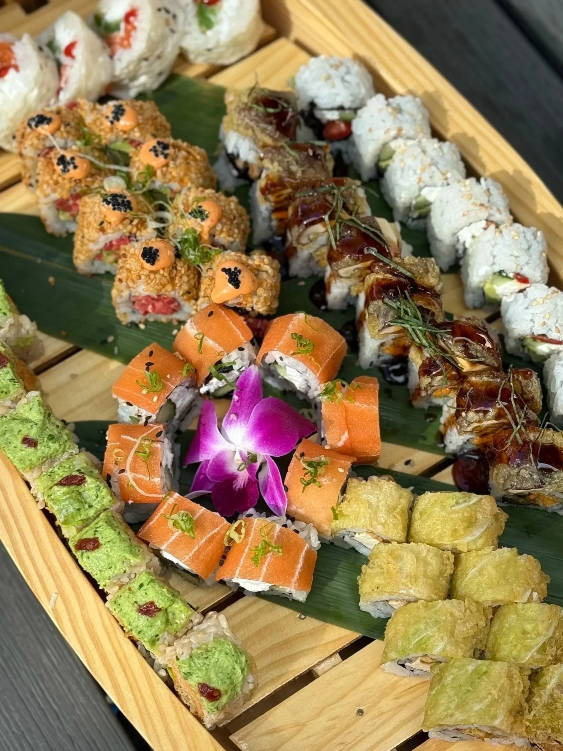 A variety of sushi rolls on a wooden platter with a pink flower and green leaves, including rolls topped with sesame and black sesame seeds, drizzled with sauces, and garnished with greens.