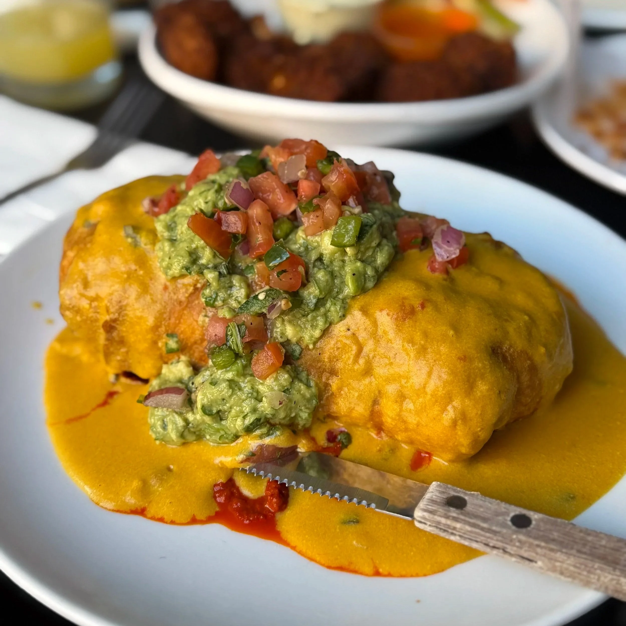 Close-up of a plate of a vegan chimichanga topped with guacamole and pico de gallo. In the background, there are bowls with cauliflower wings and some other dishes.