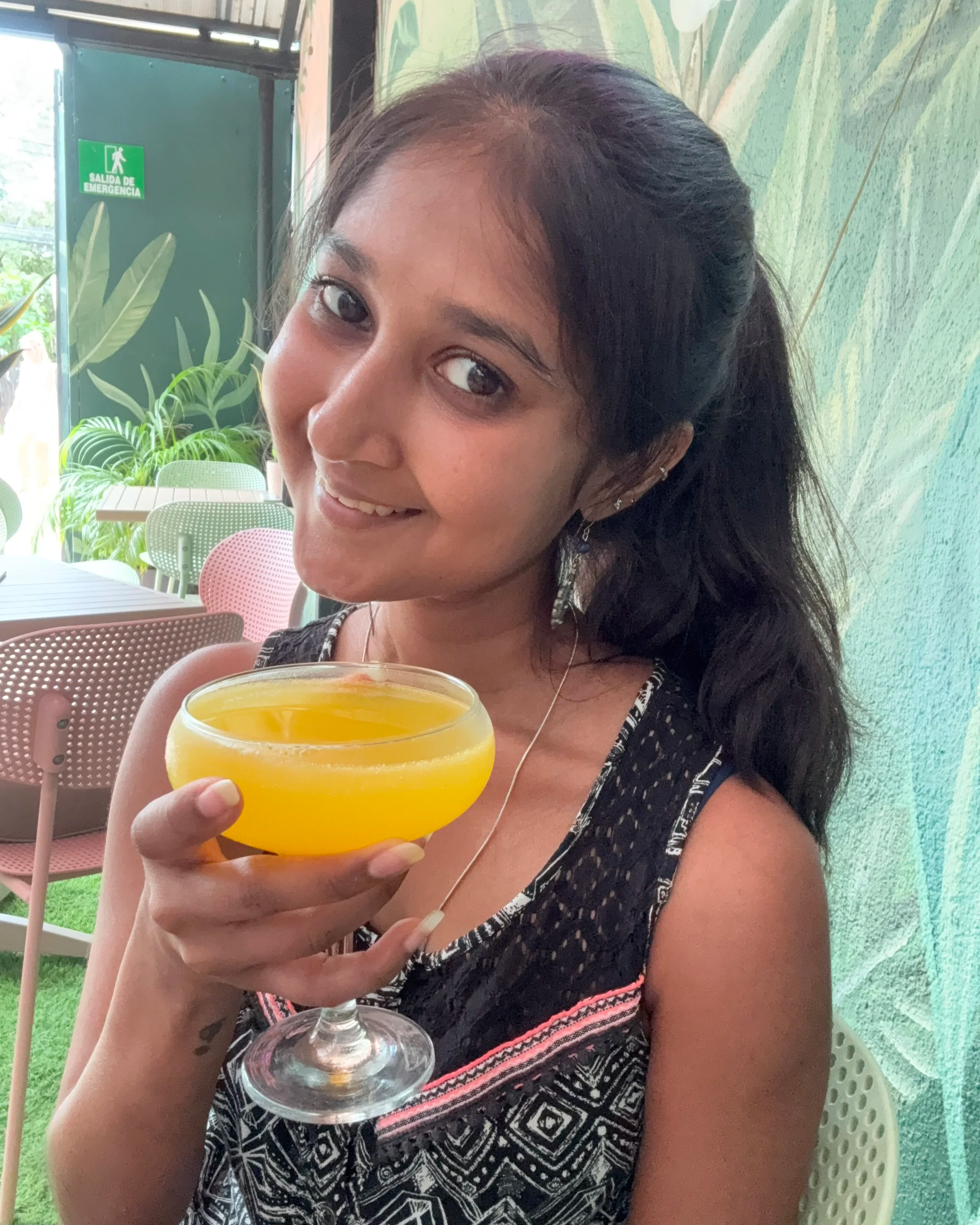 A woman holding a yellow cocktail in a glass with a stem, smiling indoors near a green wall and colorful chairs in the background.